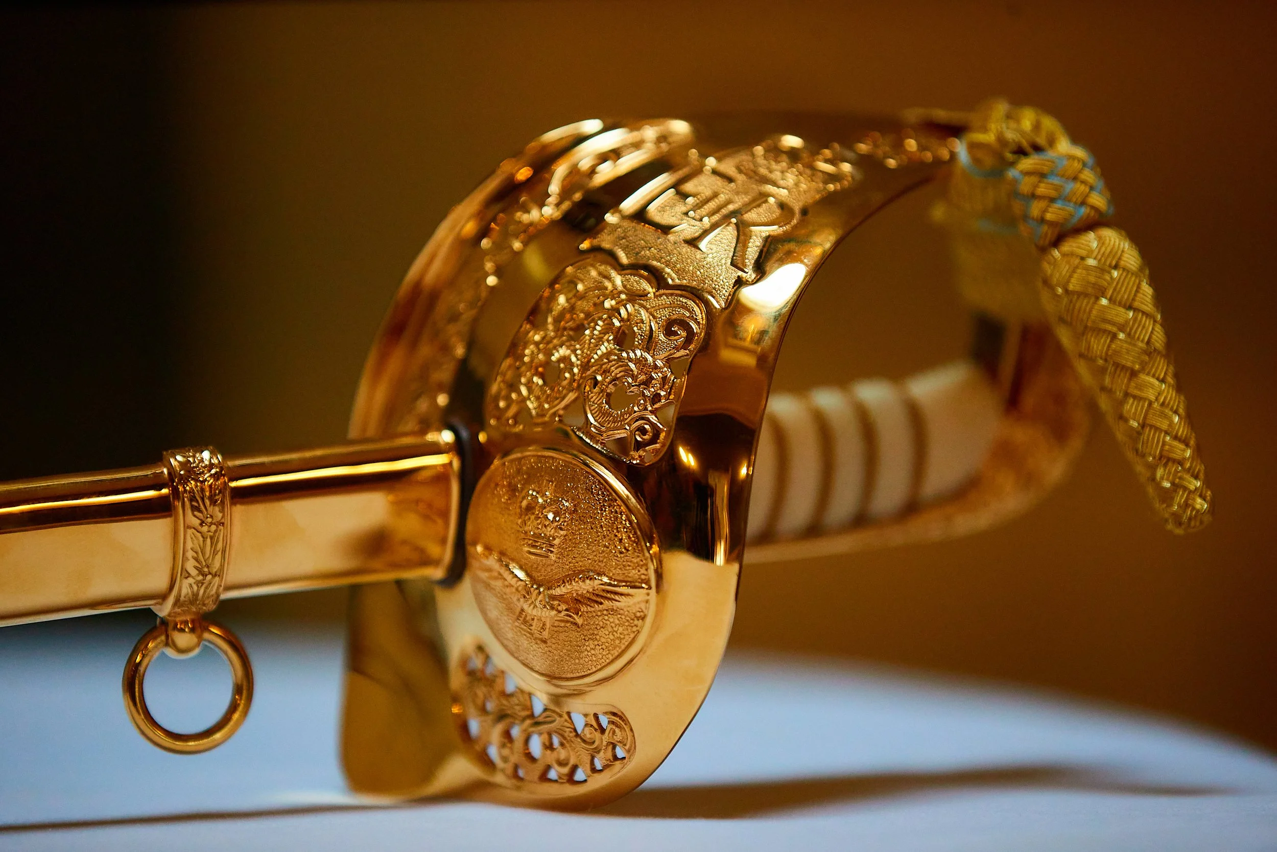 Close-up of a gold-colored decorative brooch with intricate lace-like designs, featuring a crown emblem and a small loop, attached to a woven fabric band.