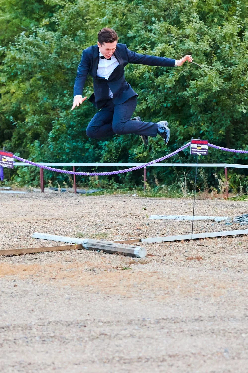A man in a business suit jumping over a barrier on a gravel path with green foliage in the background.