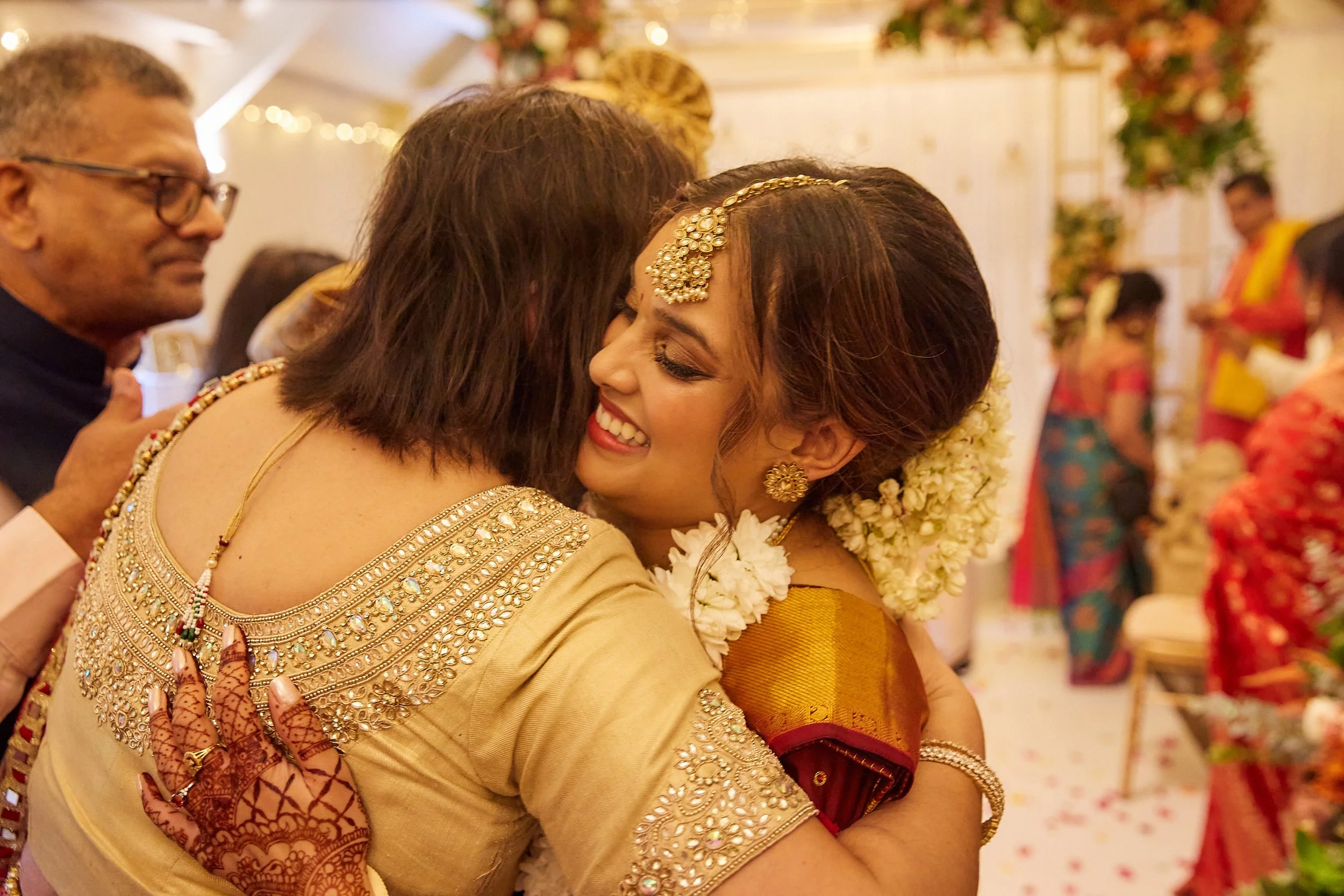 A woman with traditional jewelry and flowers in her hair is hugging another woman at a festive celebration. Both are smiling warmly, surrounded by other people, some dressed in colorful traditional attire.