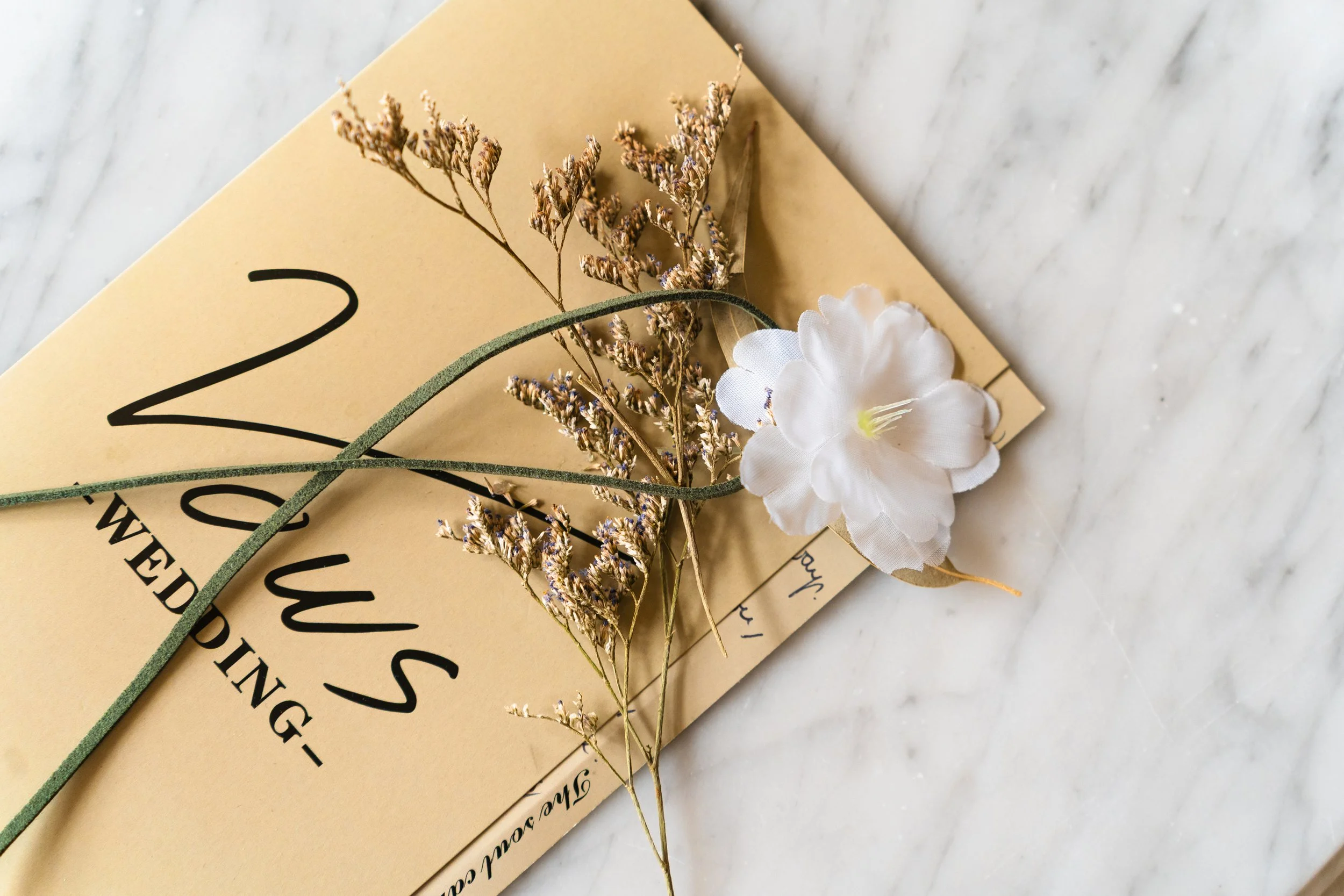 A beige wedding invitation with black text, decorated with a small bouquet of dried lavender, green suede ribbon, and a white artificial flower, placed on a marble surface.