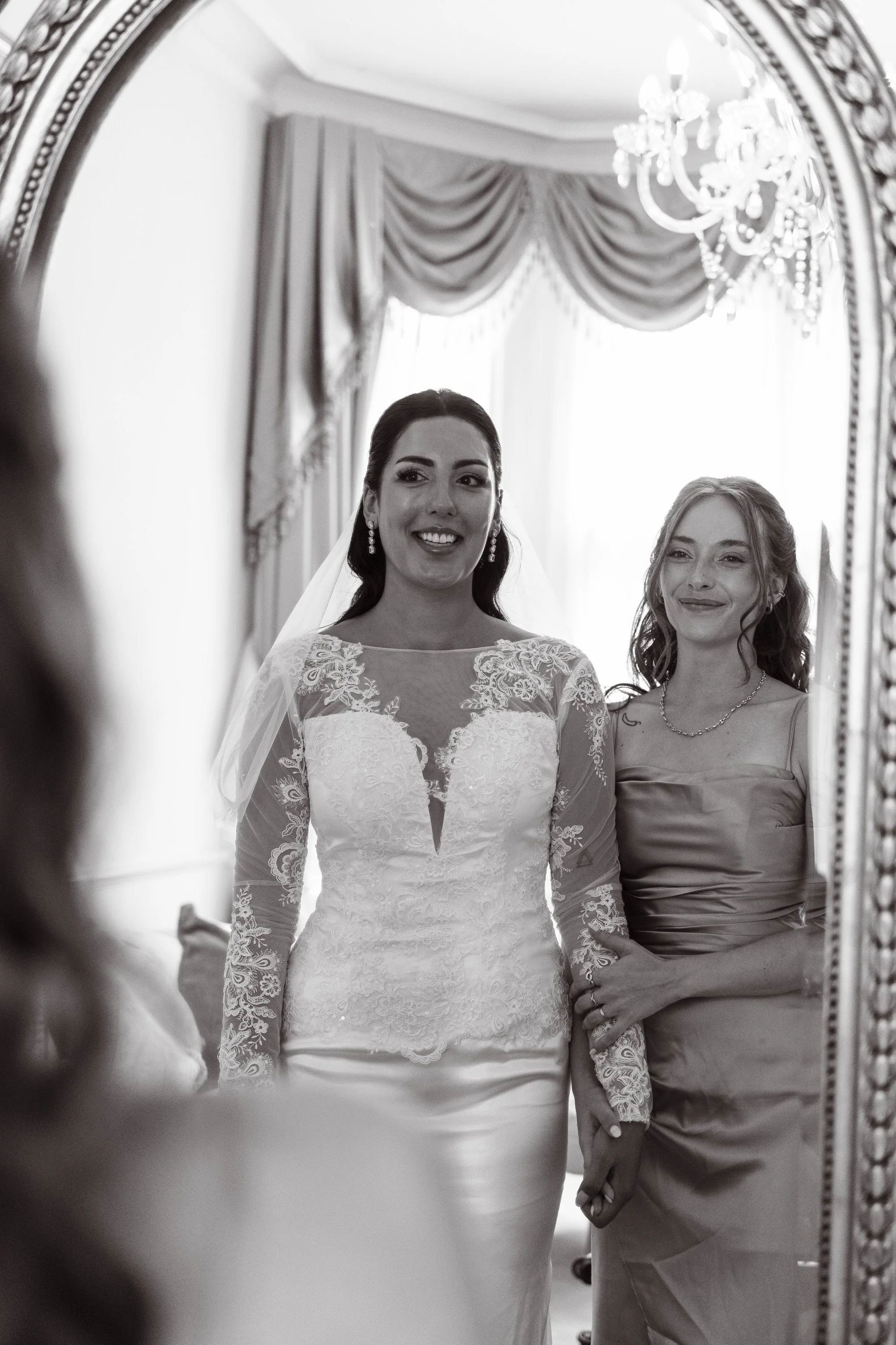 A bride in a wedding dress looking at her reflection in a mirror, smiling, with a woman beside her (possibly her mother) in a satin dress, holding her hand, in a room with elegant curtains and a chandelier.