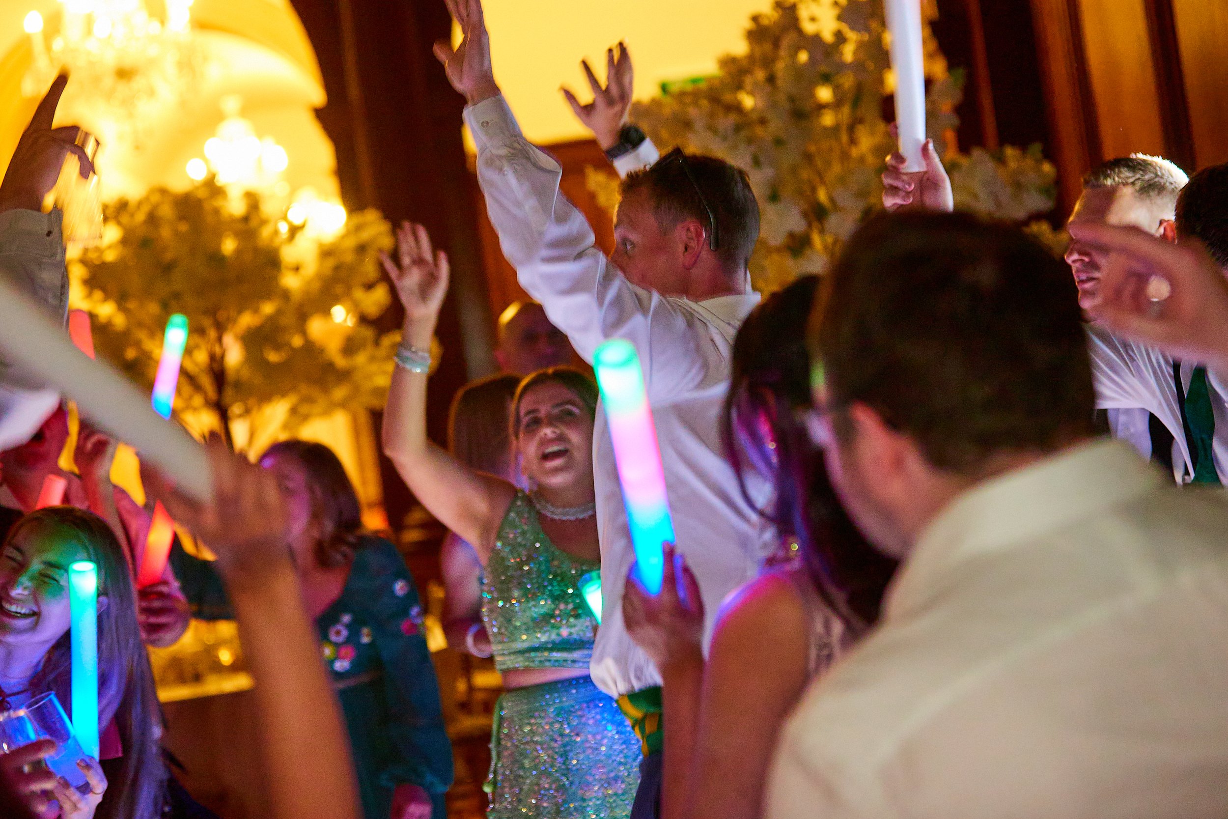 People dancing and celebrating at a party, with colorful glow sticks and warm indoor lighting.
