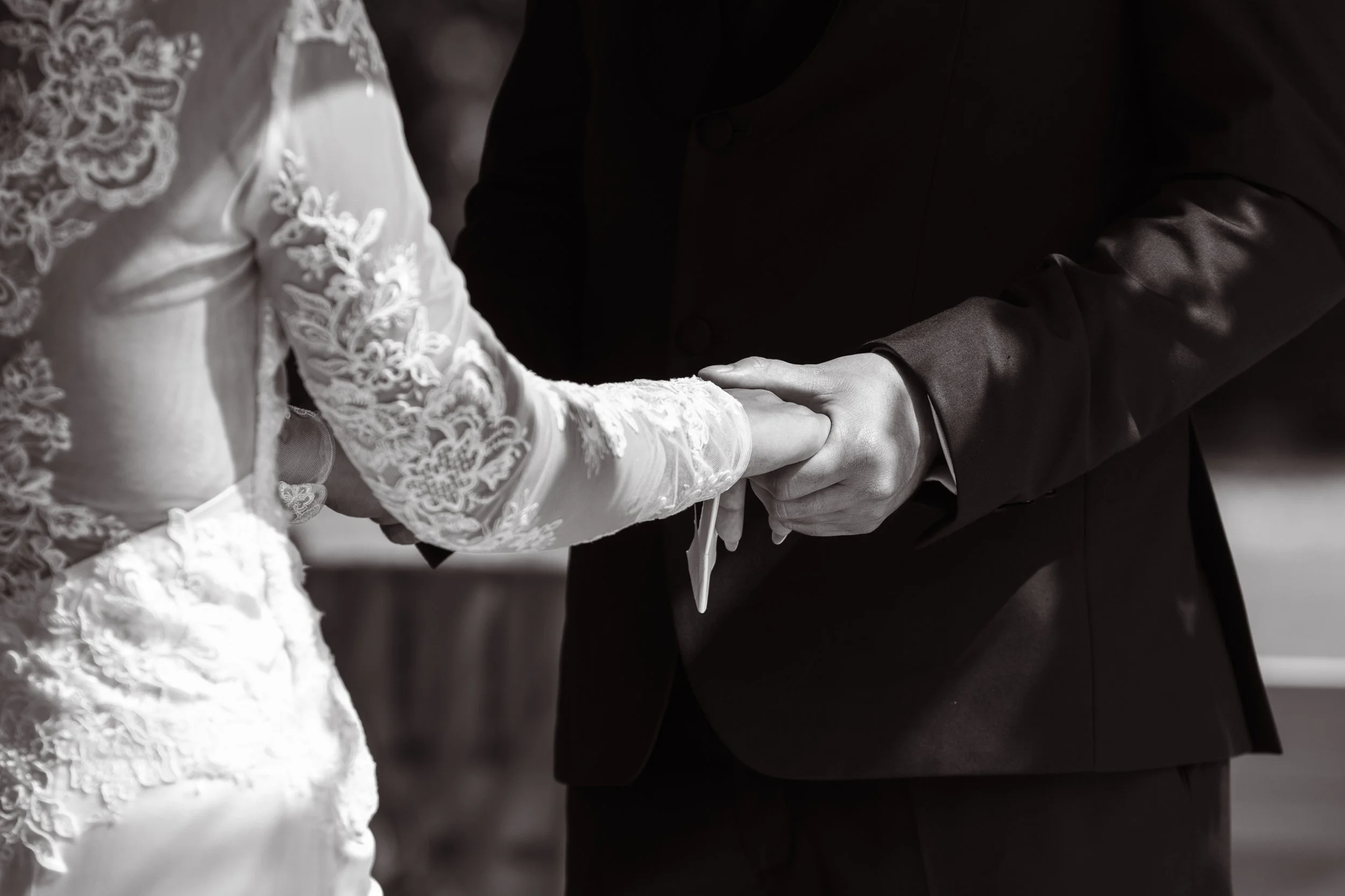 A bride and groom holding hands during their wedding ceremony. The bride is wearing a lace wedding dress and the groom is in a dark suit.