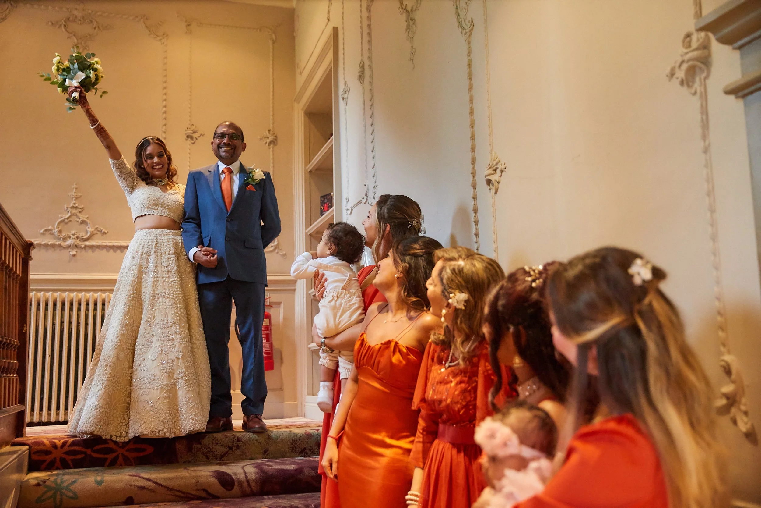A wedding scene with the bride and groom standing on a staircase, holding hands. The bride is wearing a white lace wedding dress and holding a bouquet, while the groom is dressed in a blue suit with an orange tie. Several women, dressed in orange and