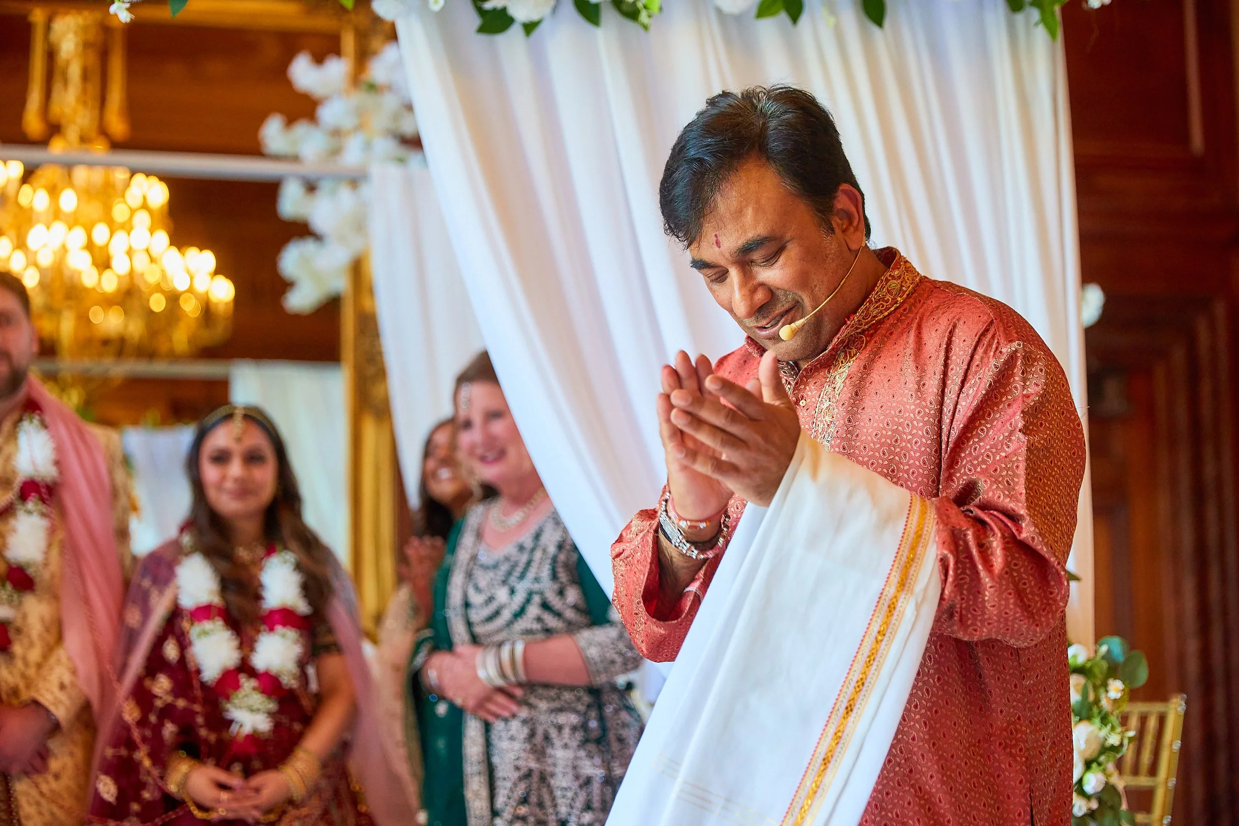 A man in traditional Indian attire with his hands pressed together in prayer during a wedding ceremony, surrounded by smiling women and a decorated venue with flowers and drapes.