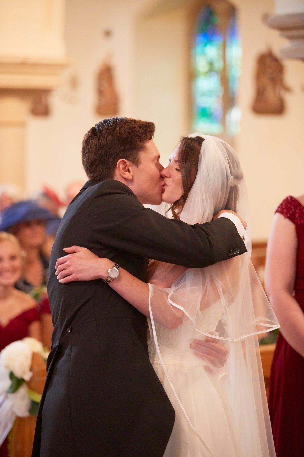 A bride and groom kissing during their wedding ceremony inside a church with stained glass windows; guests are visible in the background.