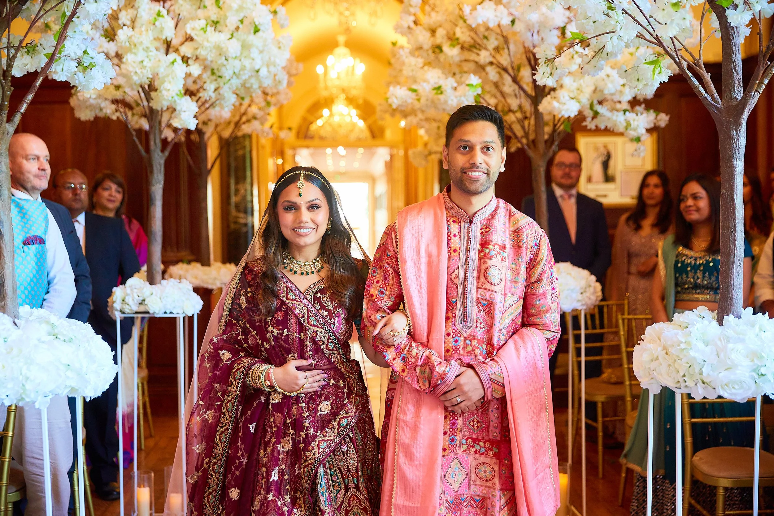 A happy couple in traditional Indian wedding attire standing arm-in-arm in a decorated ceremony room with guests in the background, surrounded by white floral arrangements and trees with white blossoms.