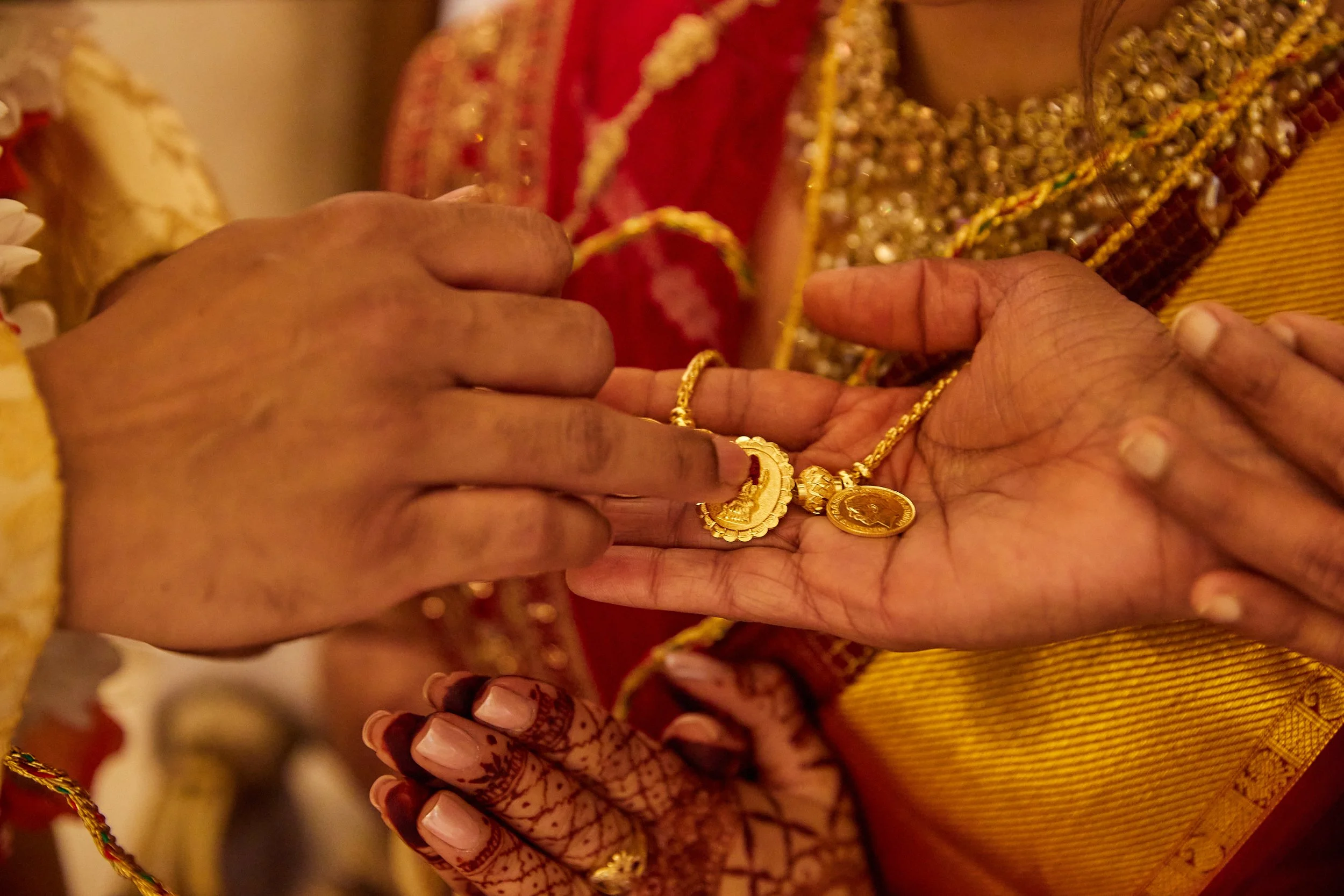 Close-up of hands exchanging gold jewelry, including necklaces with pendants, during a traditional Indian ceremony. The participants are dressed in colorful, embroidered traditional attire.