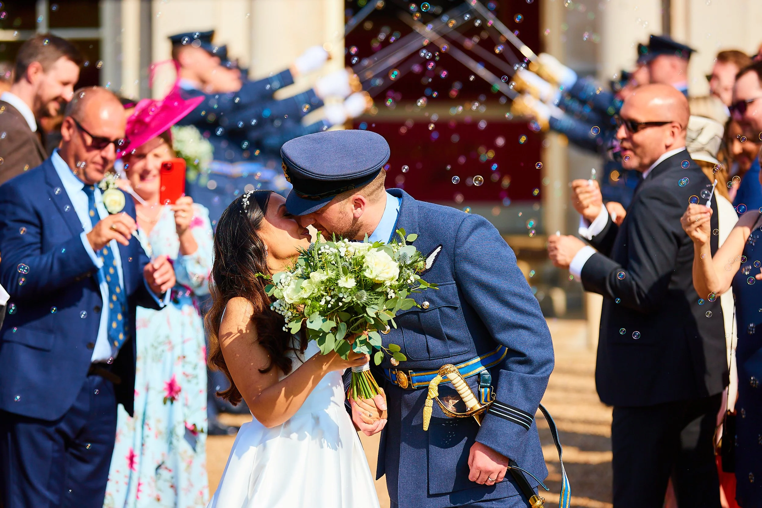 A bride and groom in military uniforms kissing during their wedding surrounded by friends and family celebrating outside, with confetti in the air.