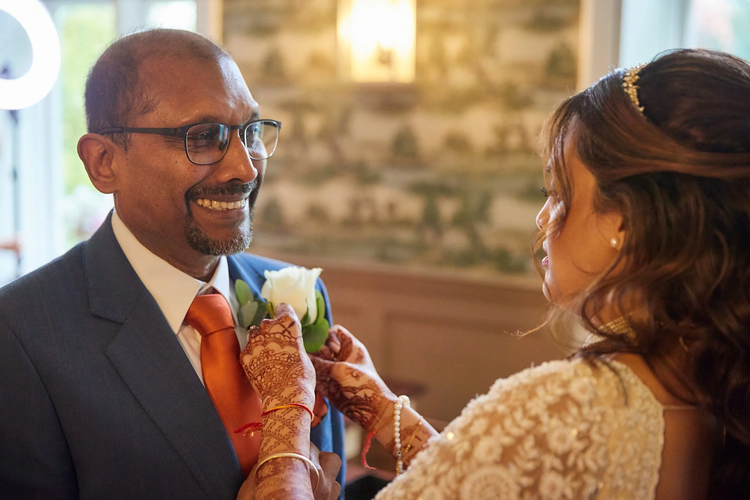 A happy man in a suit with glasses, orange tie, and a white rose boutonniere is having a moment with a woman dressed in a white lace outfit. The woman is adjusting the man's boutonniere. They are indoors with a warm background.