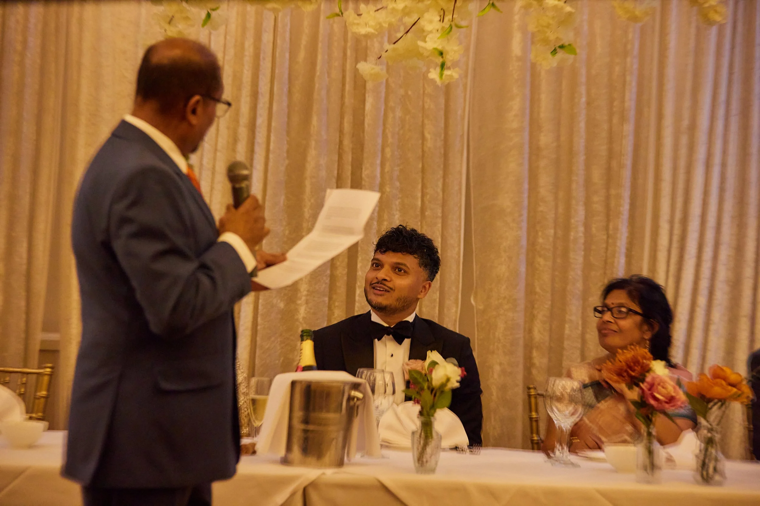 Man in suit giving a speech at a formal event with two guests listening, decorated with flowers
