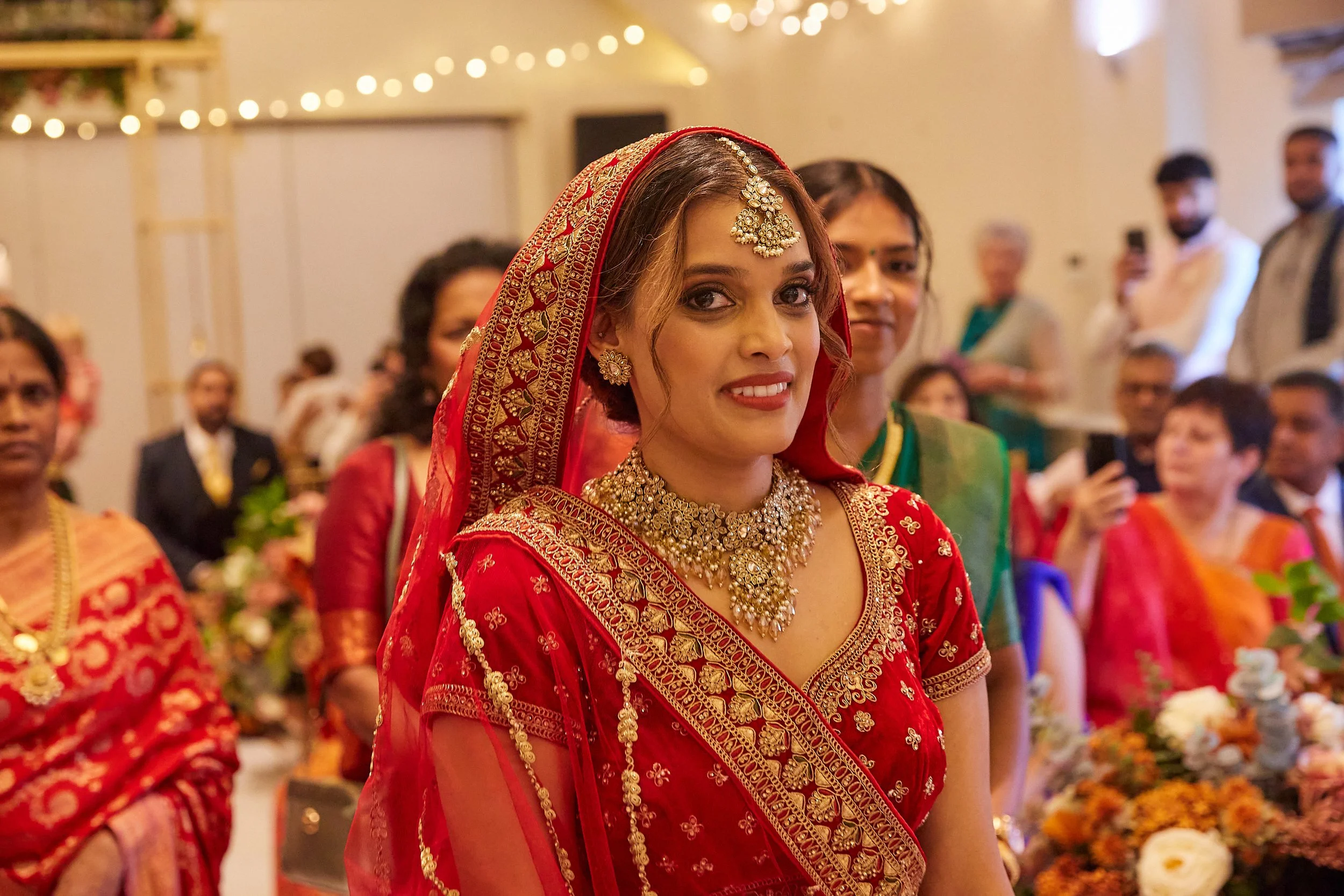 A woman in a red traditional Indian outfit and jewelry, smiling at a celebration, surrounded by others in colorful attire.