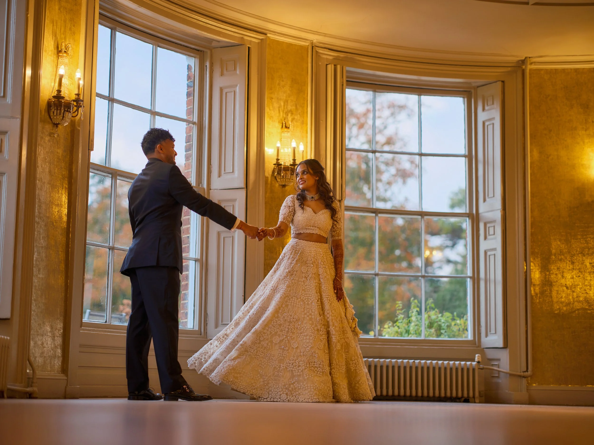 A bride and groom holding hands in a warmly lit room with large windows and autumn trees outside.