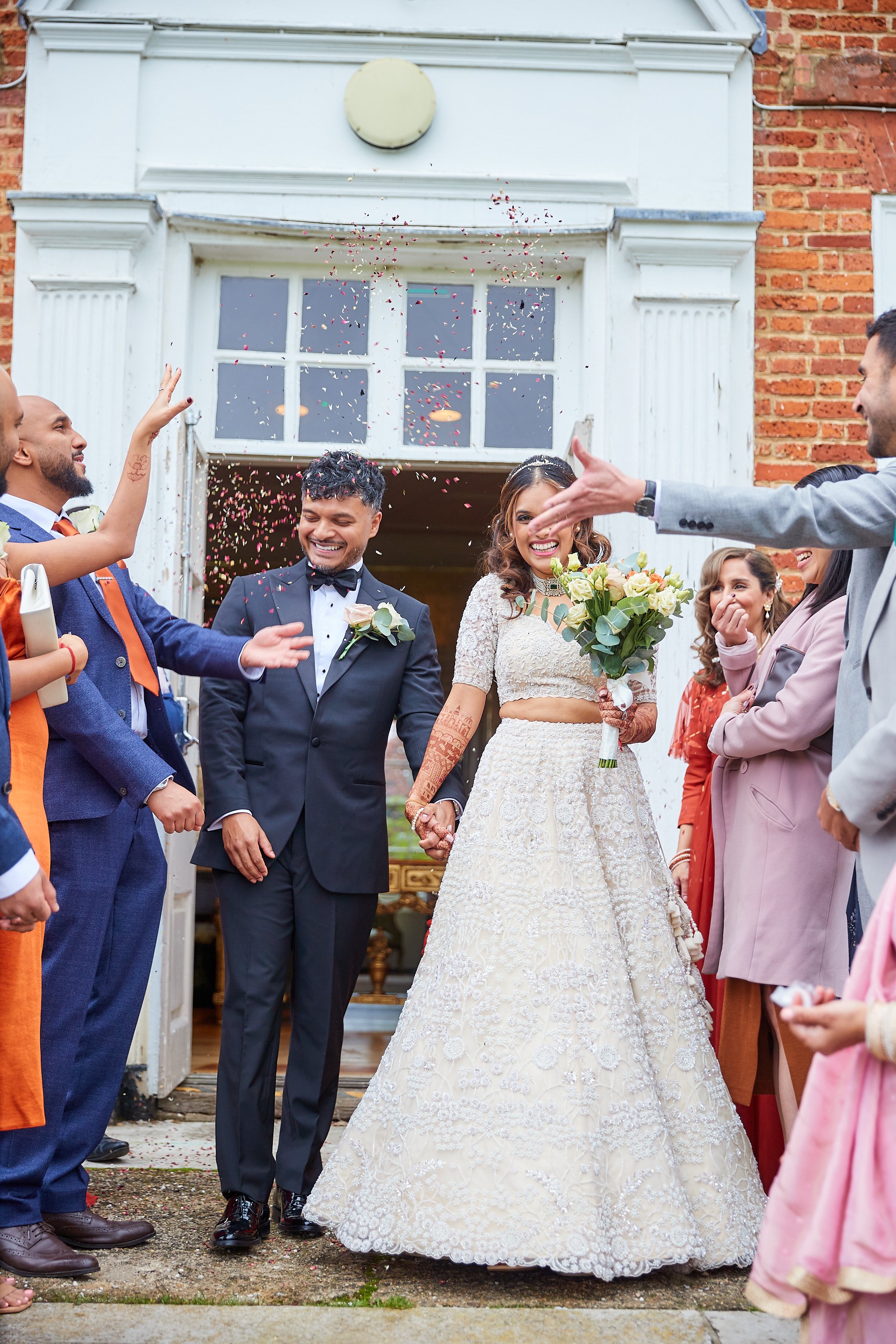 Couple leaving wedding venue, surrounded by friends and family, with confetti falling, bride holding bouquet, groom in tuxedo, wedding dress with intricate lace, diverse group celebrating.