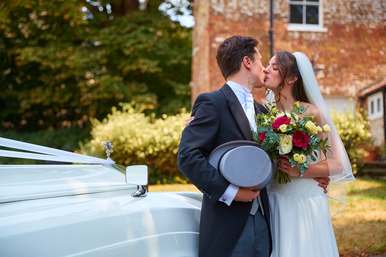 A bride and groom sharing a kiss outdoors on their wedding day, with the bride holding a bouquet of red and yellow roses, and the groom holding a gray hat and a gray fedora, standing next to a white vintage car with a London-style side mirror.