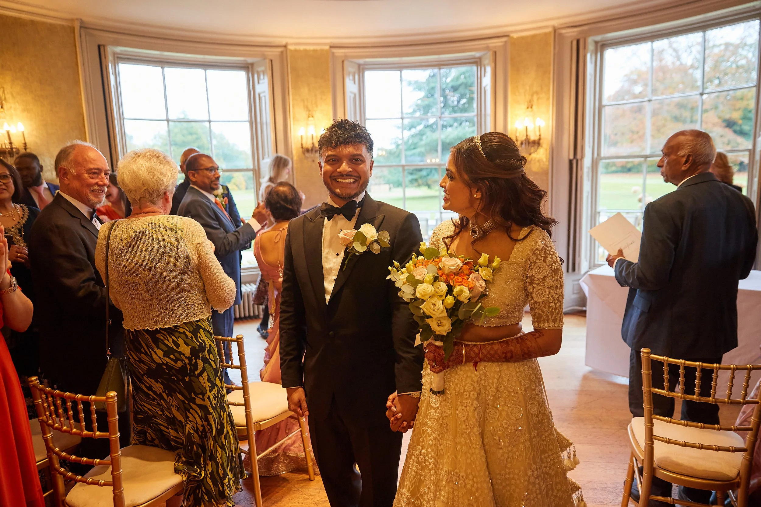 Bride and groom holding hands at their wedding celebration, surrounded by family and friends in a decorated indoor venue with large windows.