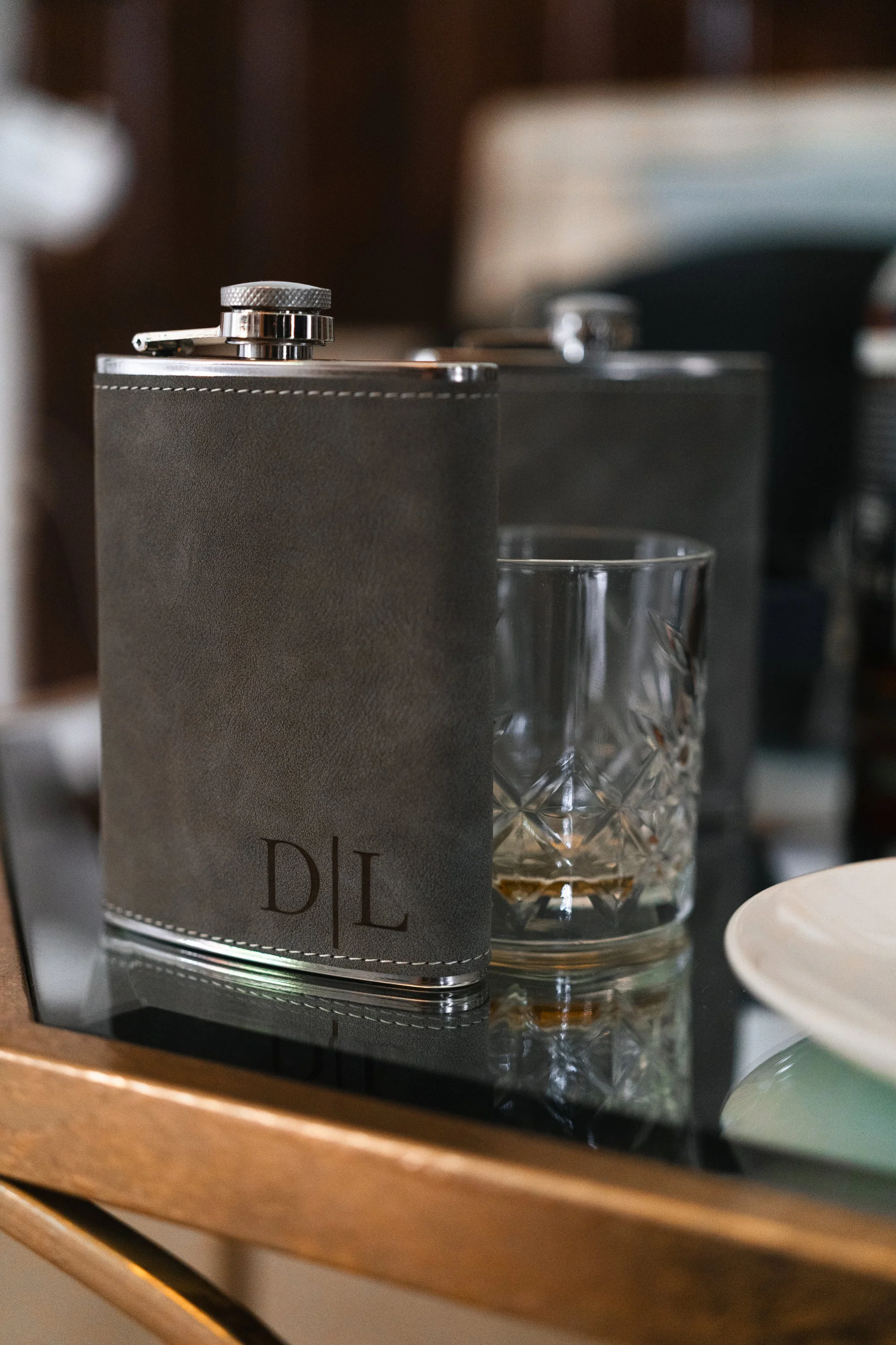 A metal flask with the initials DIL and a glass of whiskey on a reflective surface, with another similar flask in the background.