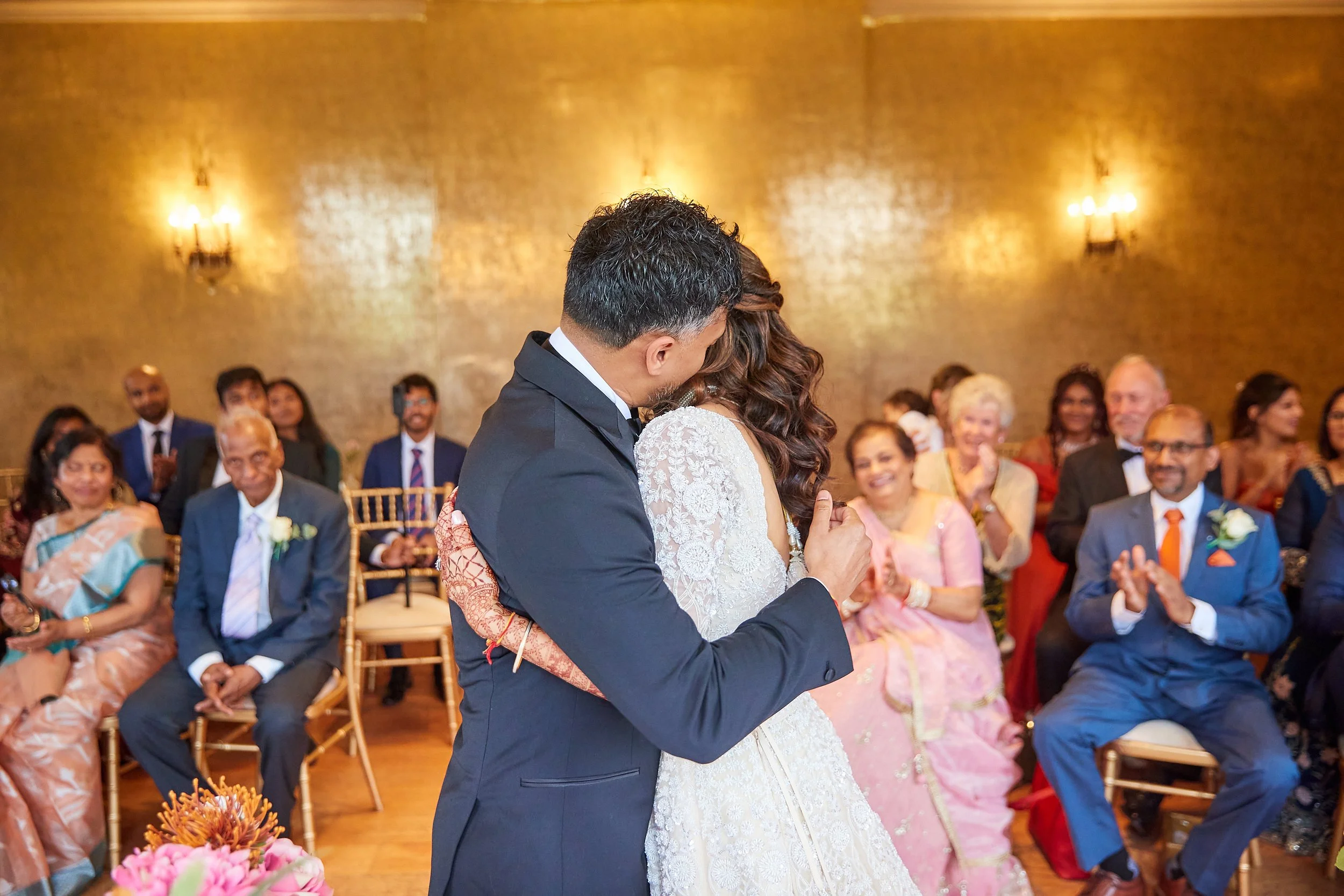 A bride and groom dance closely during their wedding ceremony, surrounded by seated guests clapping and smiling in a warmly lit venue with a gold-toned brick wall in the background.