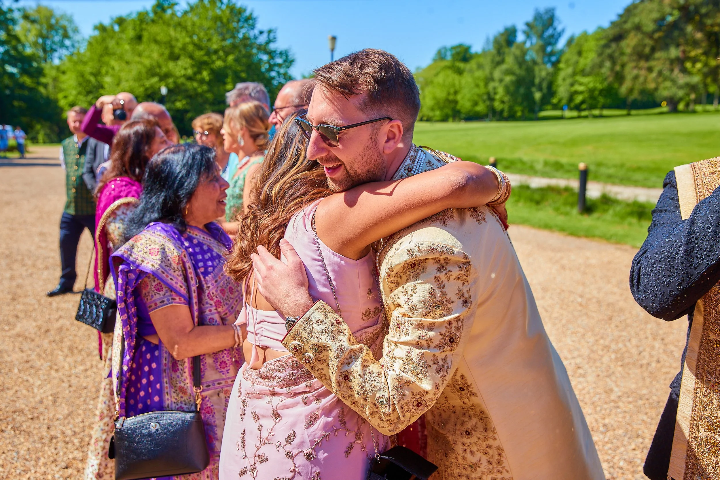 A man and woman in traditional South Asian attire embrace each other during a celebration outdoors on a sunny day. Other people in colorful clothing stand in line behind them, smiling and taking photos on a grassy field with trees in the background.