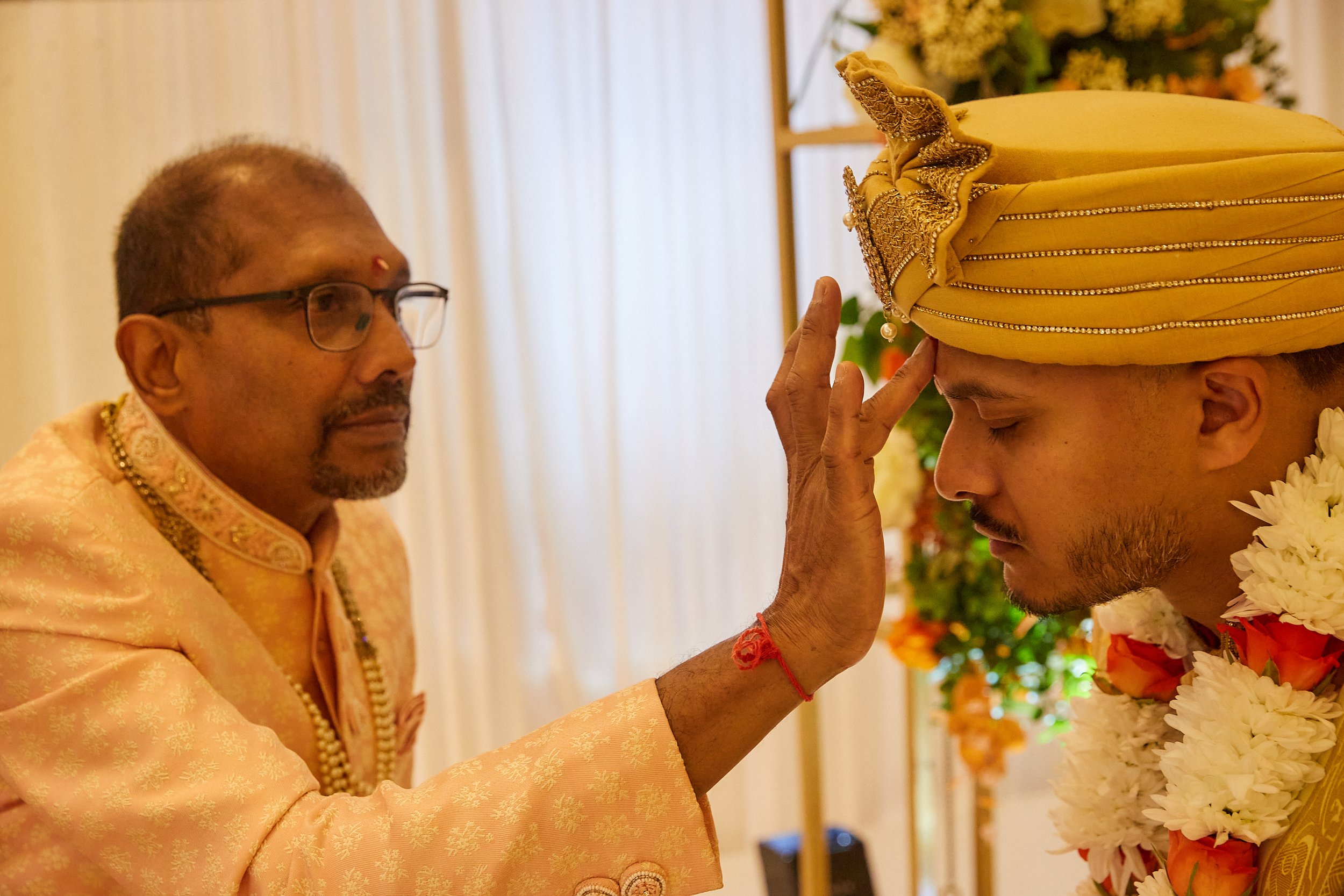 A traditional Indian wedding ceremony where a priest performs a ritual by placing his hand on the groom's forehead. The groom is wearing a yellow turban and floral garland, and the priest is dressed in a peach-colored traditional outfit.