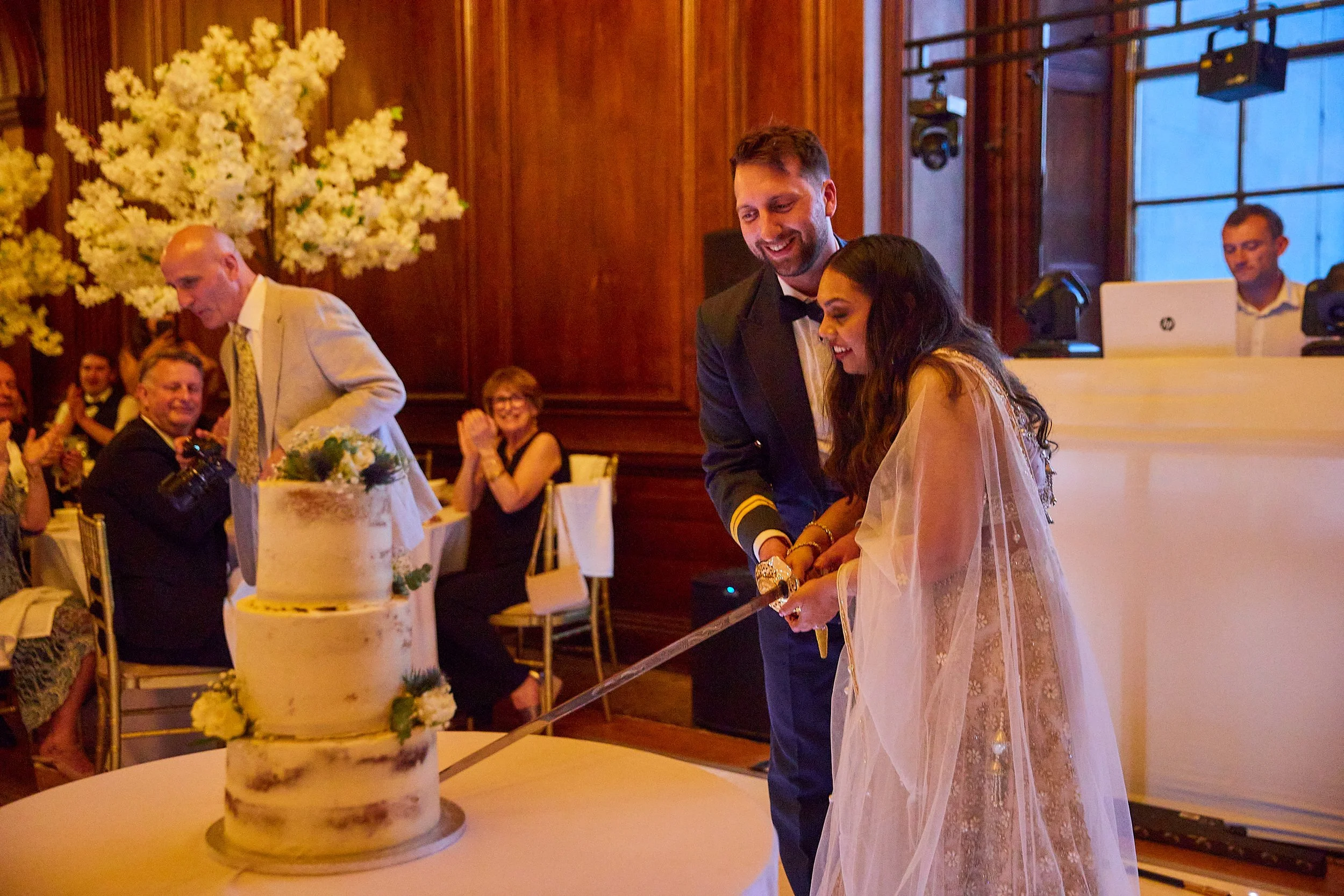 A bride and groom cut a wedding cake together, surrounded by guests at a formal wedding reception, with a large floral arrangement in the background.