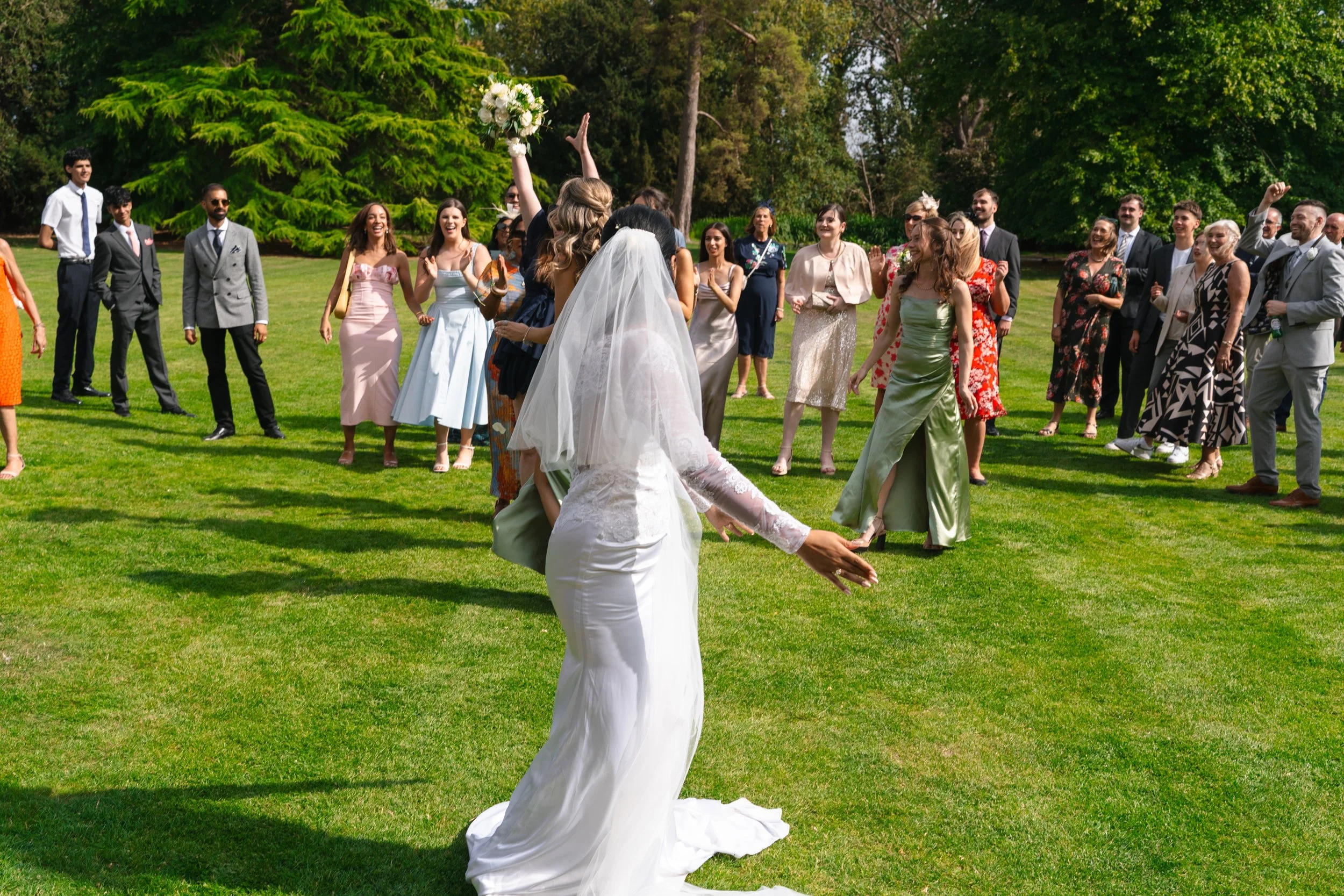 A bride in a white wedding dress and veil is throwing her bouquet to a group of guests on a green lawn. The guests, dressed in colorful dresses and suits, are smiling and reaching out to catch the bouquet, with trees and blue sky in the background.