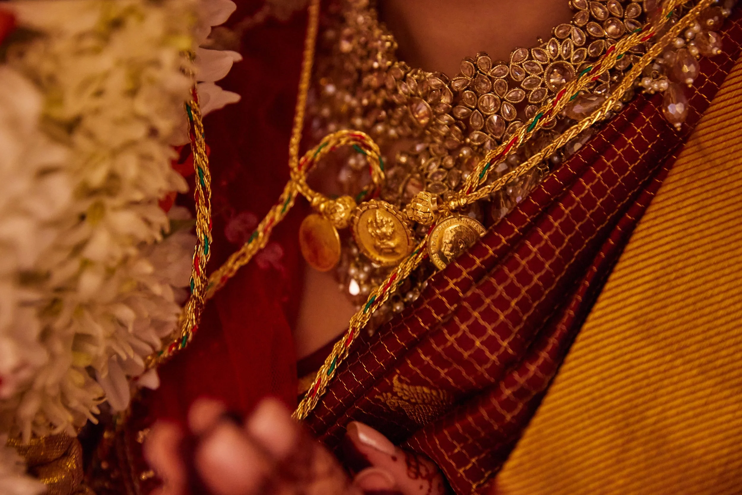 Close-up of a woman wearing multiple traditional gold jewelry pieces, including necklaces with coin pendants, layered over a red and gold patterned sari, with a floral garland partially visible on the left side.