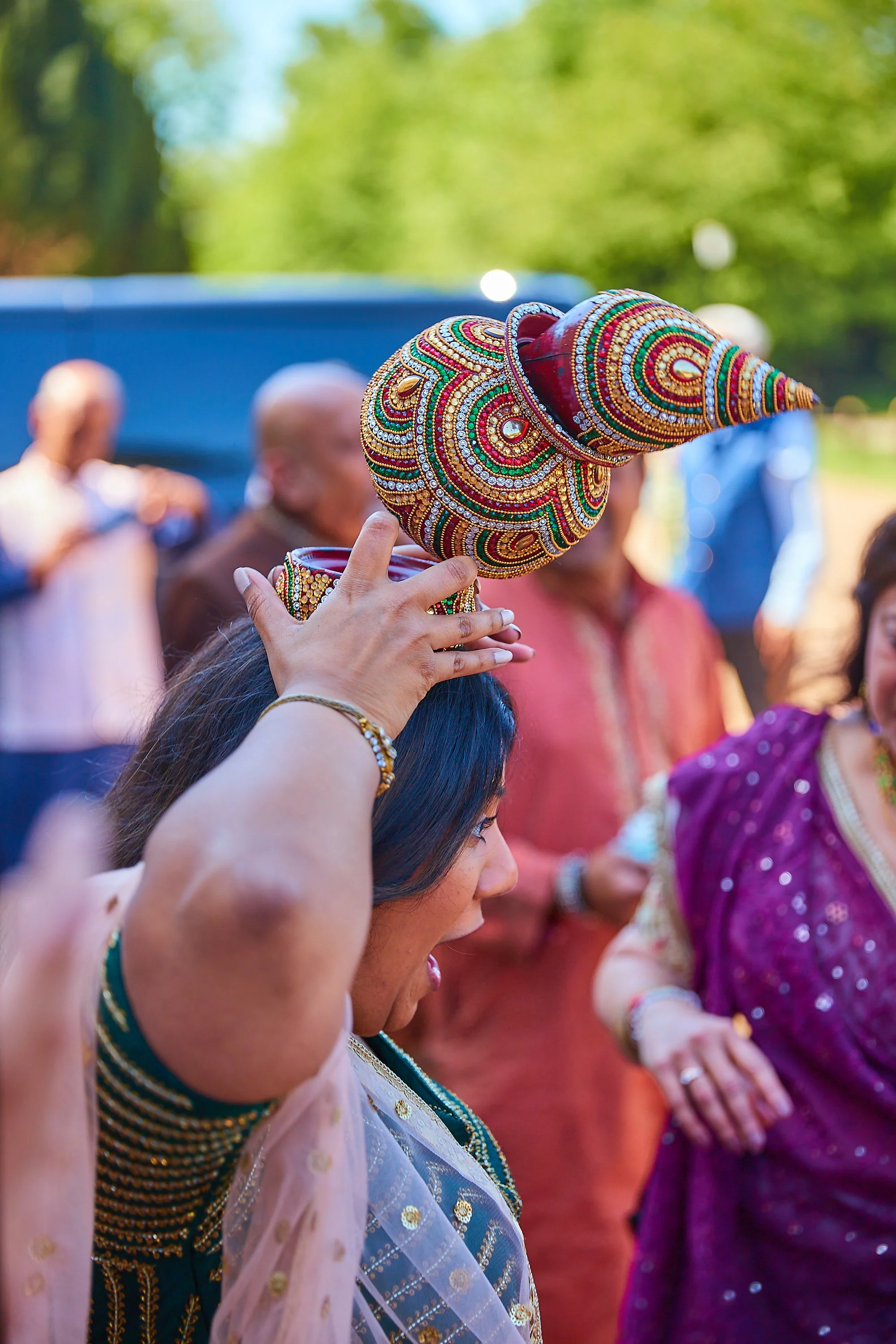A woman wearing traditional Indian attire is holding a large, decorated conch shell above her head during a celebration or ceremony outdoors, with other people dressed in traditional clothing in the background.