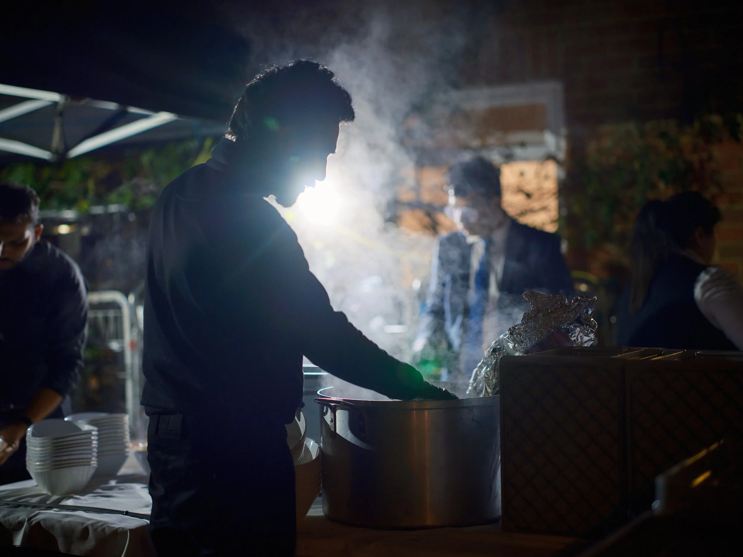 People serving food at an outdoor event at night with steam rising from a large container, silhouetted against bright backlighting.