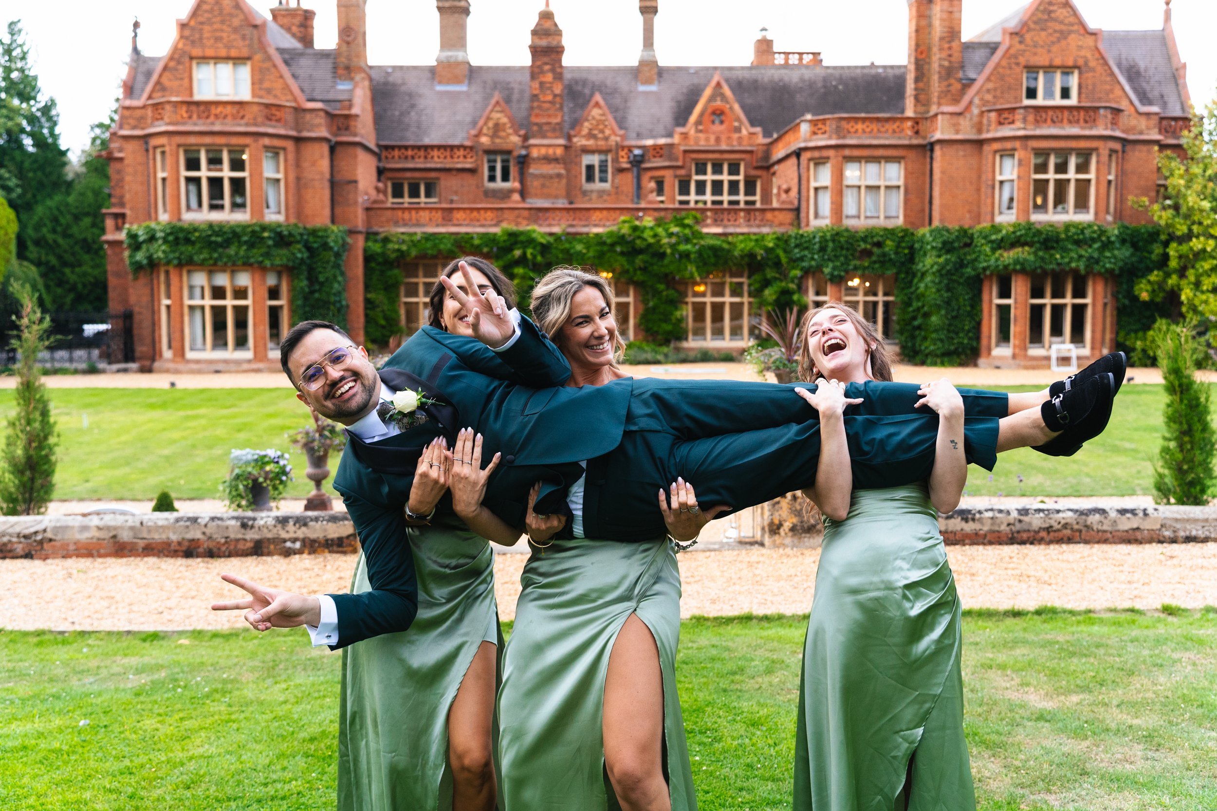 A group of people, two women in green dresses and a man in a suit, lifting a man in a suit and glasses in the air in front of a large red brick house with green climbing plants and a green lawn.