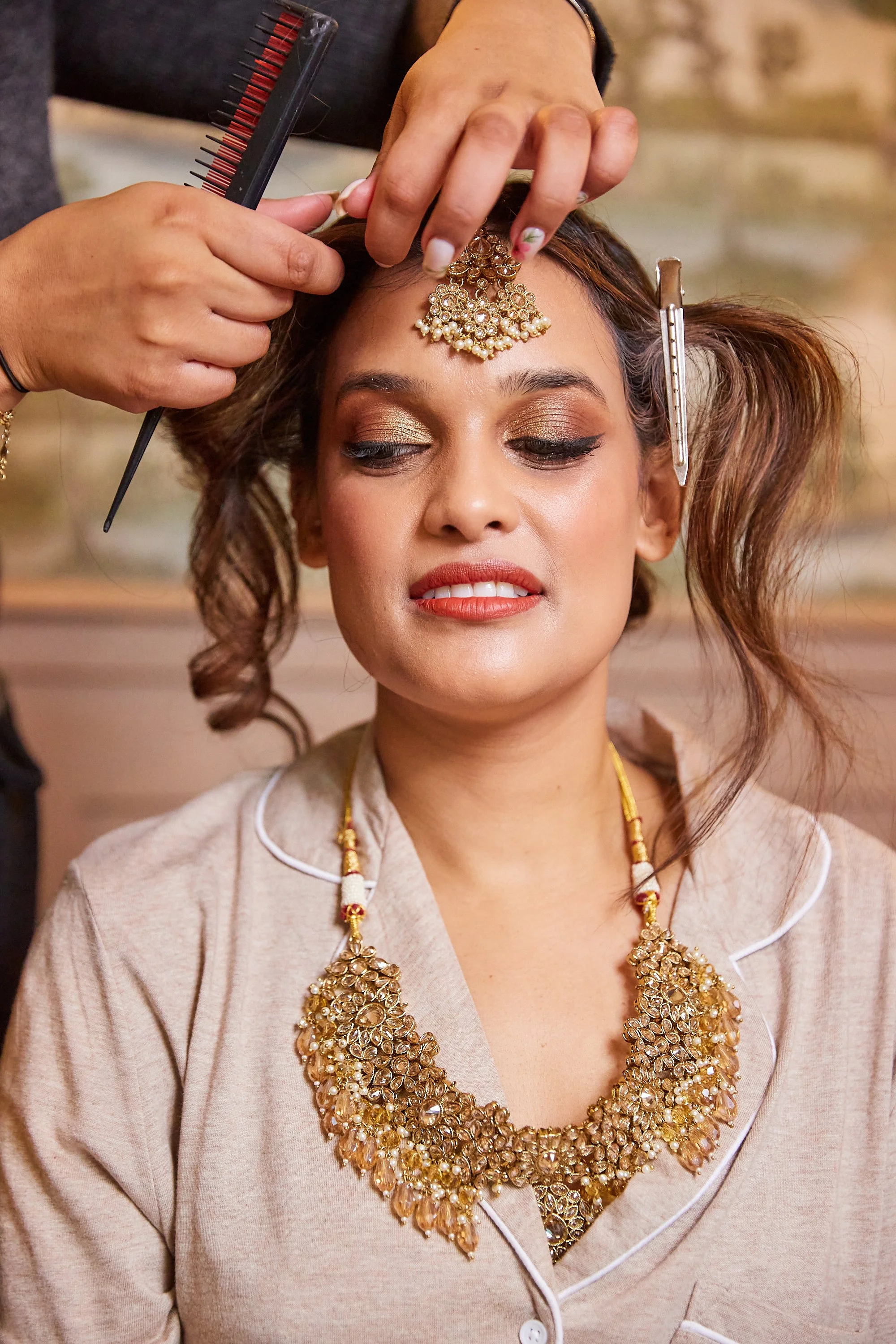 A woman with styled hair and jewelry is getting her hair and accessories arranged by a stylist. The stylist is holding a hairpin and a comb, gently adjusting the woman's hair and jewelry on her forehead as she prepares for a special occasion.