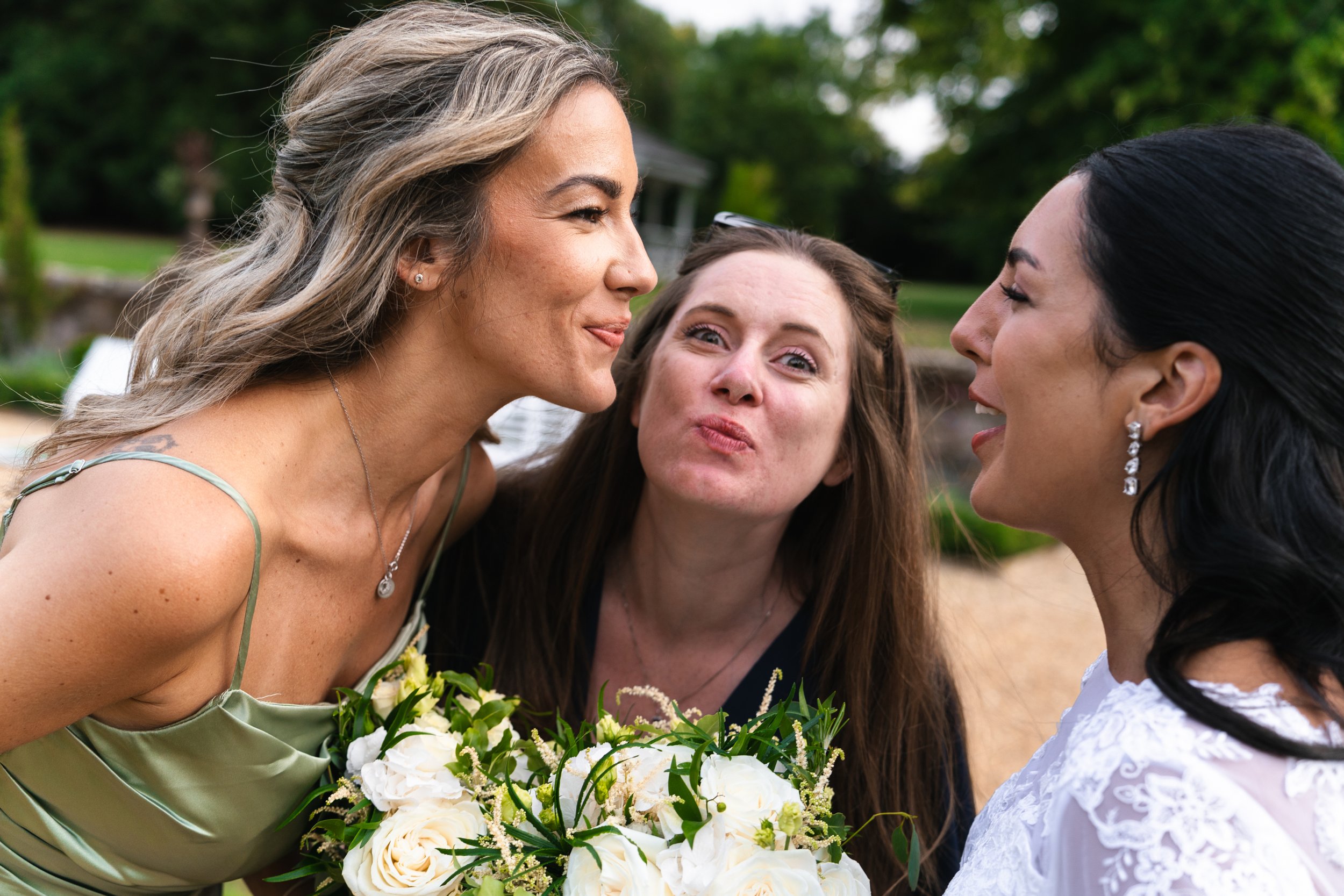 Three women are outside, making playful faces and appearing to be having fun. One woman is holding a bouquet of white flowers, and the background is green with trees.