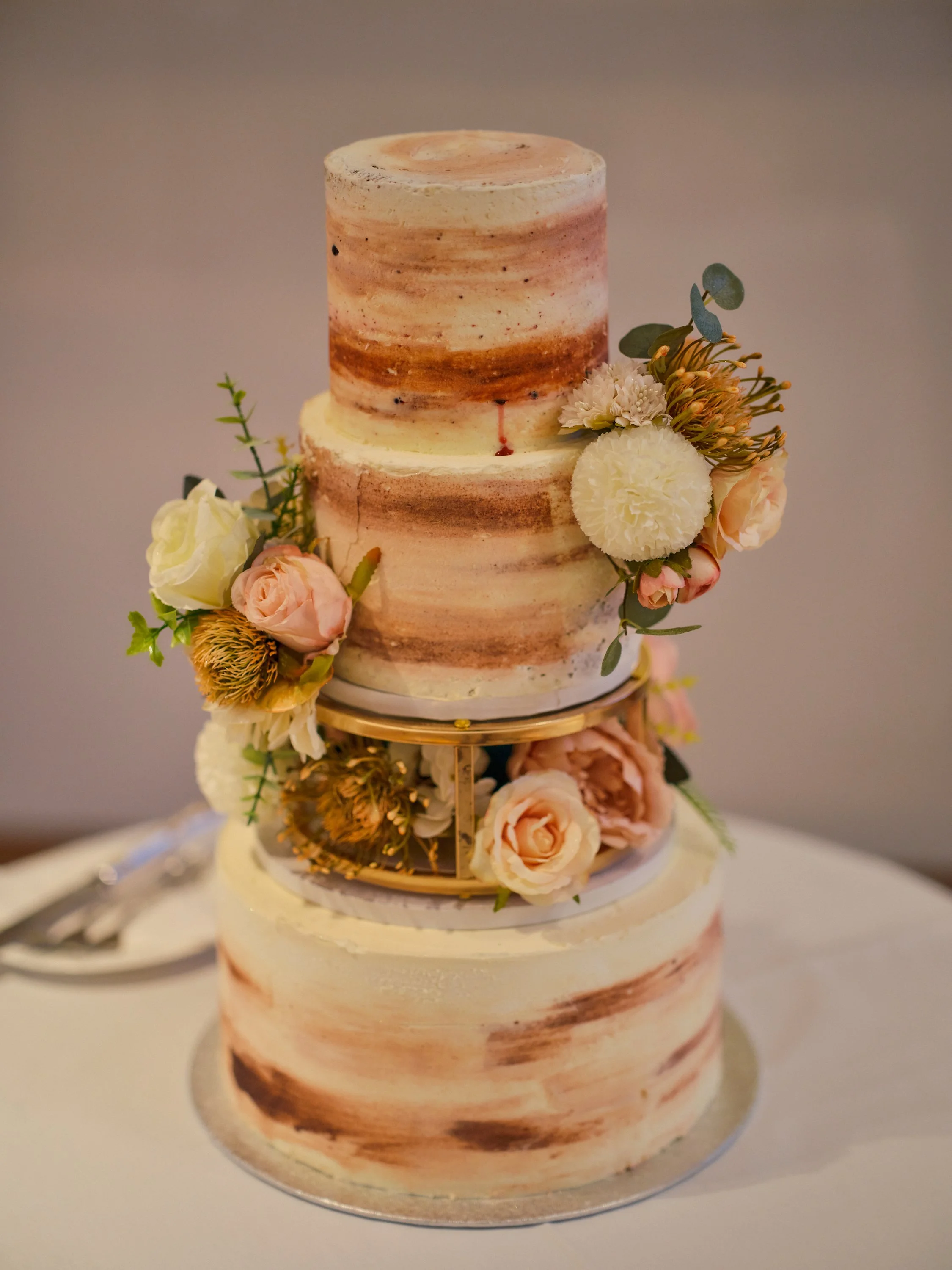 A two-tier cake with a semi-naked buttercream finish, decorated with pink and white flowers and greenery, displayed on a stand.