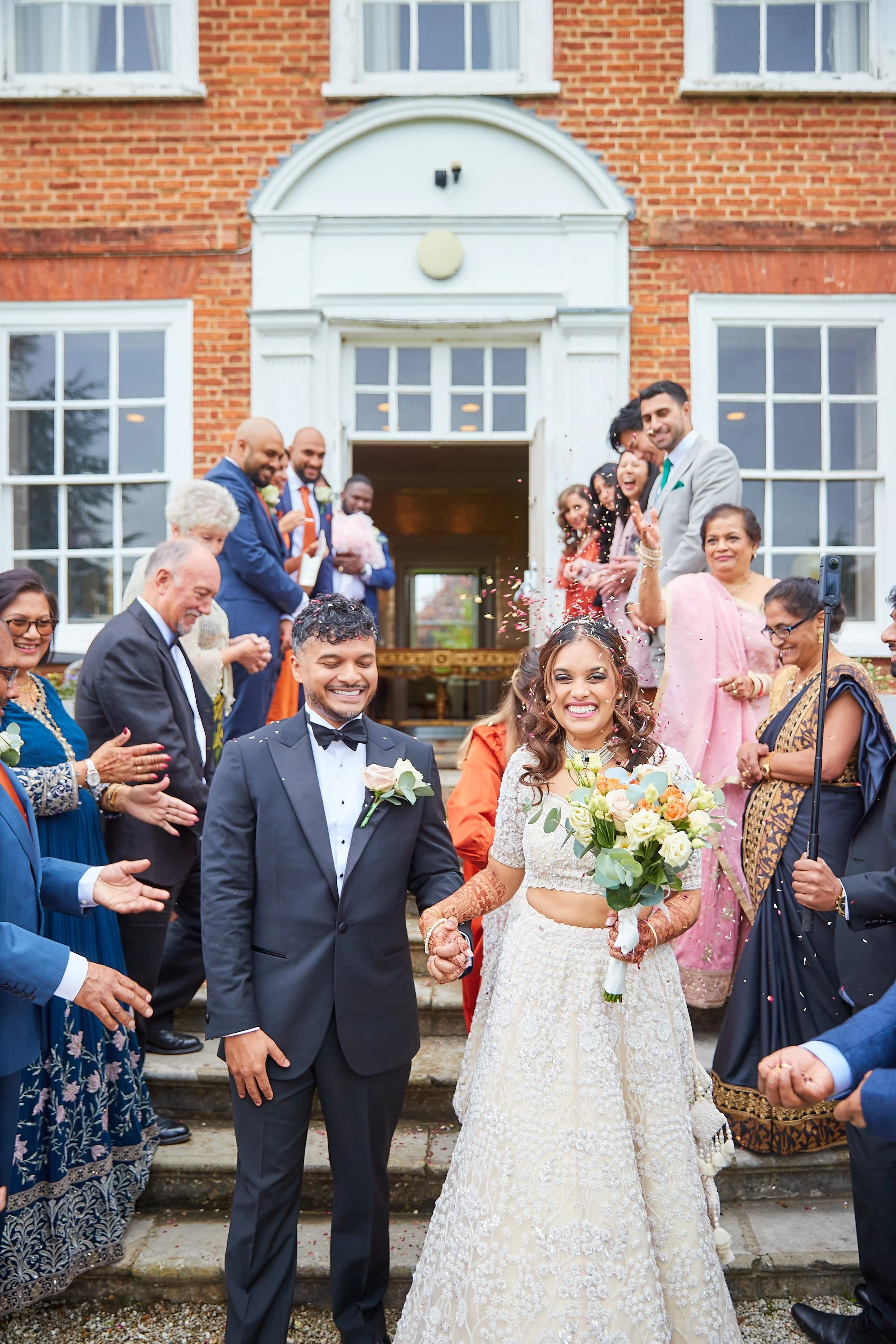 A newlywed couple walking down the steps of a brick building, celebrating with friends and family. The bride is smiling and holding a bouquet, while the groom is smiling and holding her hand. Guests are cheering and throwing confetti, with some clapp