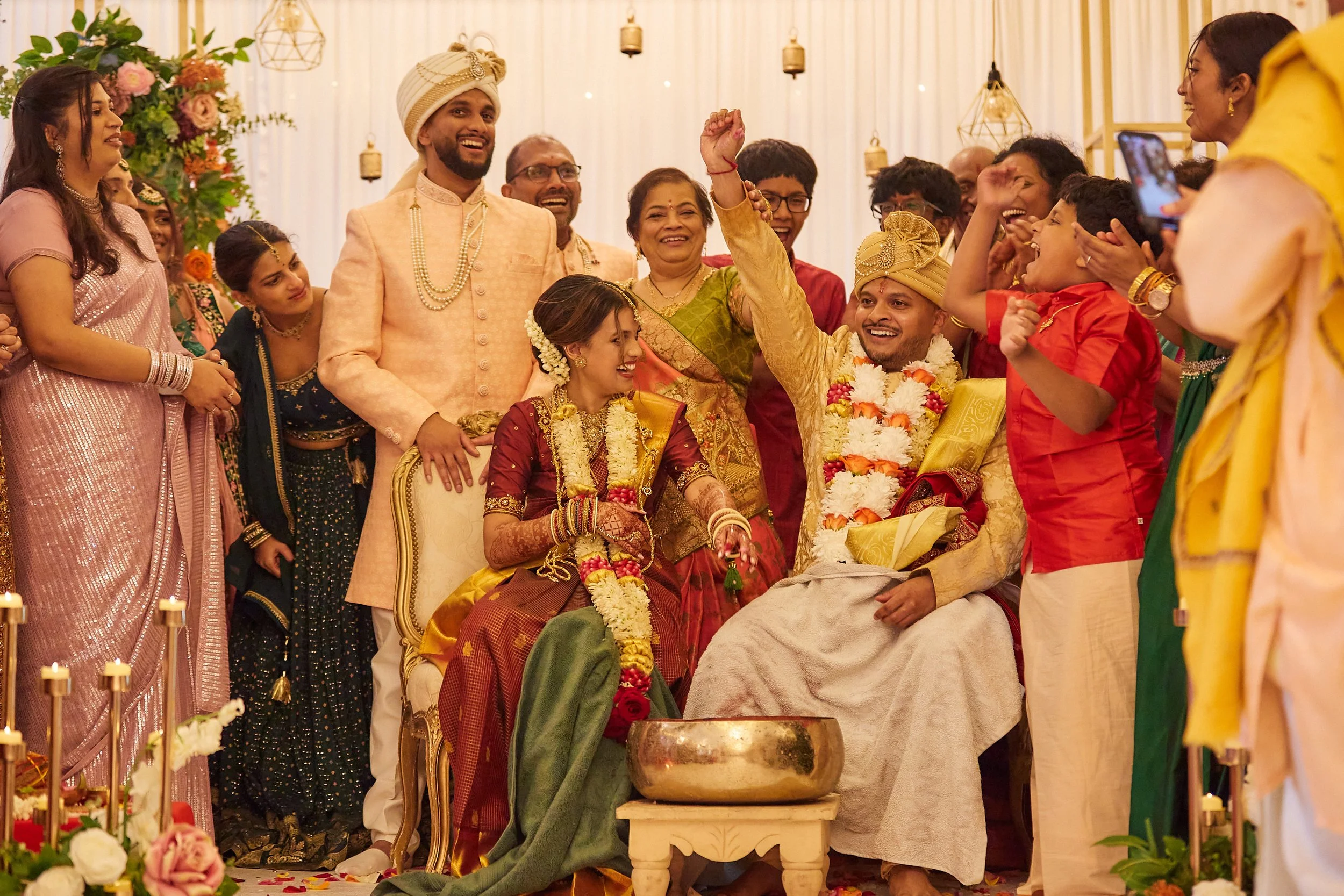 A group of Indian people celebrating a wedding, with the bride and groom sitting in the center. The bride wears a red and gold saree, and the groom wears traditional cream and gold attire with a turban. They are surrounded by family and friends, some