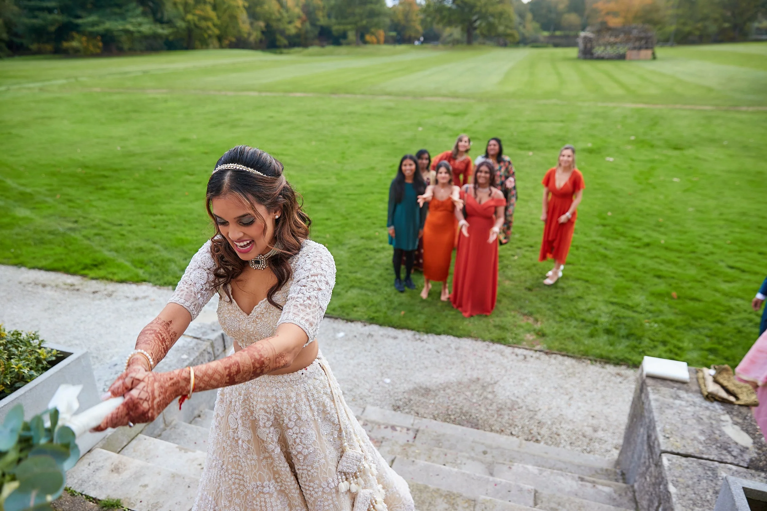 A bride in a wedding dress and mehndi on her hands smiling as she places flowers, with a group of women in colorful dresses standing behind her in a green outdoor setting.