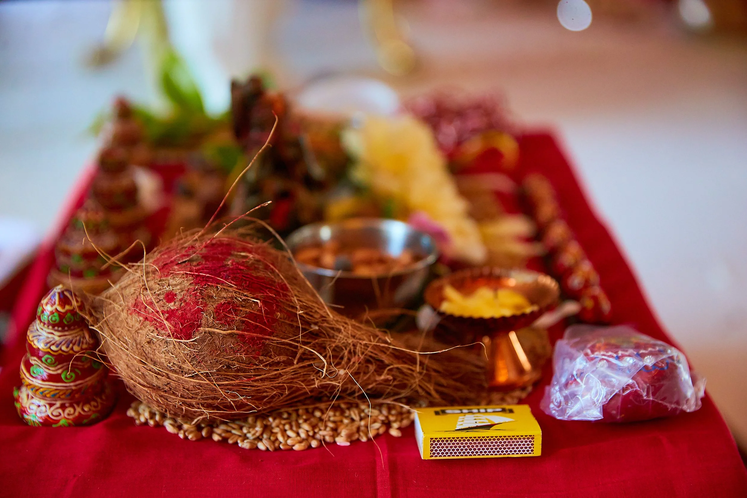 A traditional Indian festive display featuring a coconut, decorative lamp candles, a box of matches, a plastic bag, and some sweets on a red cloth.