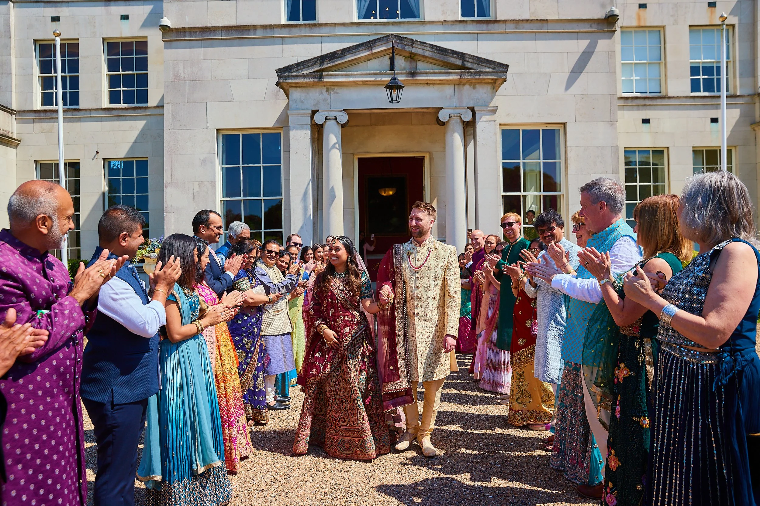 Indian bride and groom in traditional attire walking out of a building, surrounded by a crowd of people in colorful traditional clothing, celebrating outdoors.
