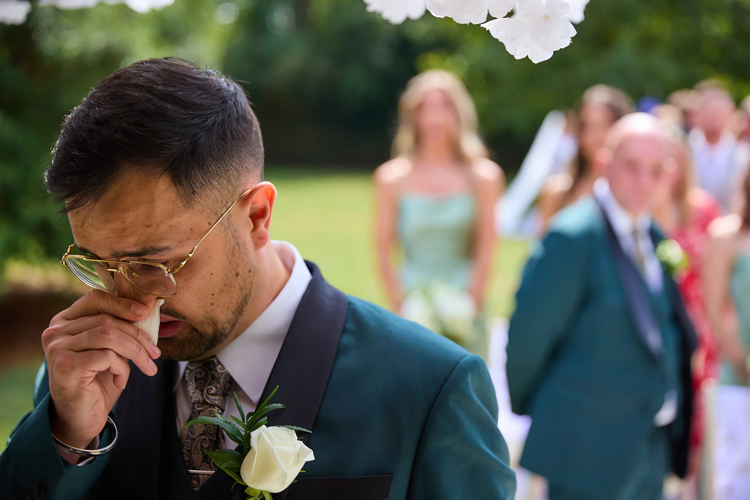A man wearing a suit and glasses, holding a white rose, with his head bowed and face touching his hand during an outdoor wedding ceremony. Blurred group of people in the background.