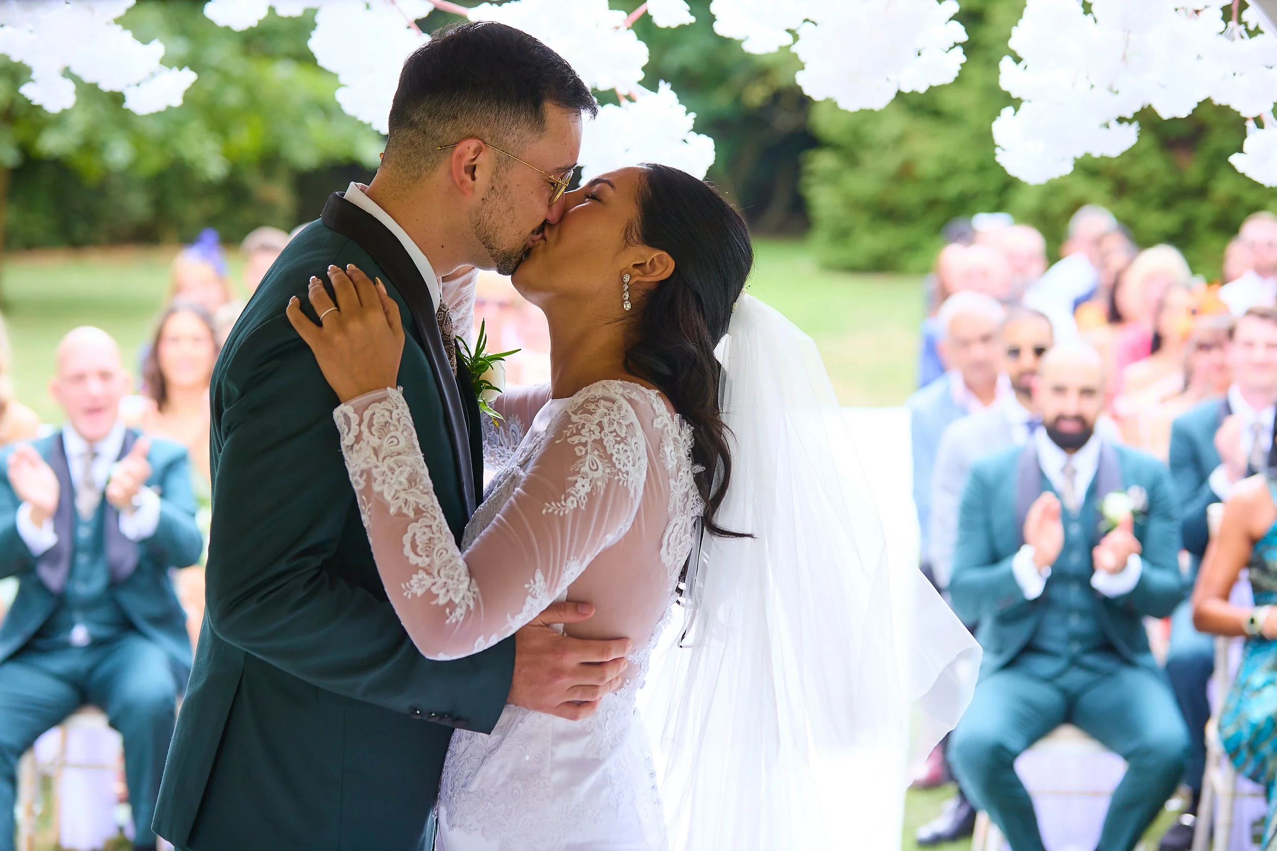 A bride and groom kiss during their outdoor wedding ceremony as guests clap in the background.