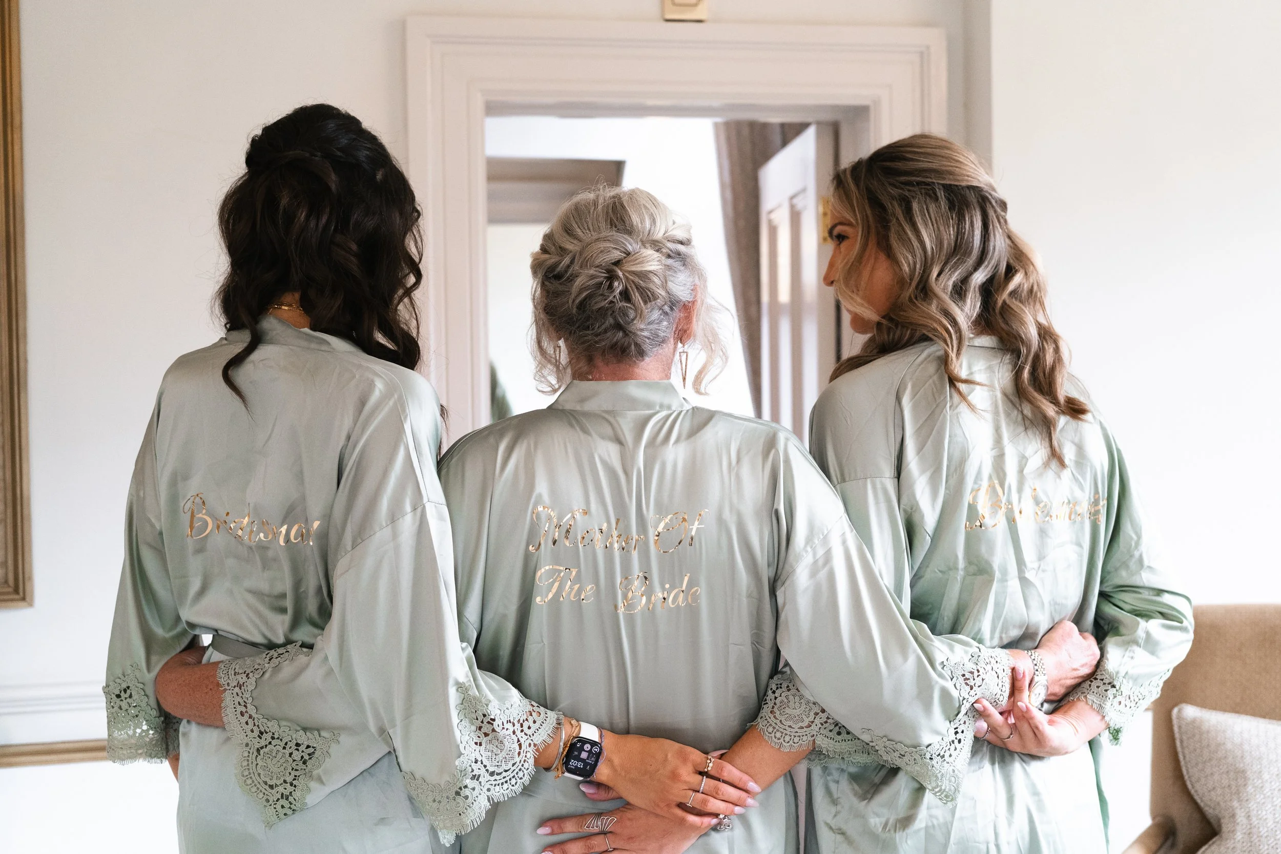 Three women wearing matching light green satin robes with lace cuffs, in a room near a mirror. The robes have titles embroidered in gold on the back: 'Bridesmaid,' 'Mother of the Bride,' and 'Groom's Sister.'