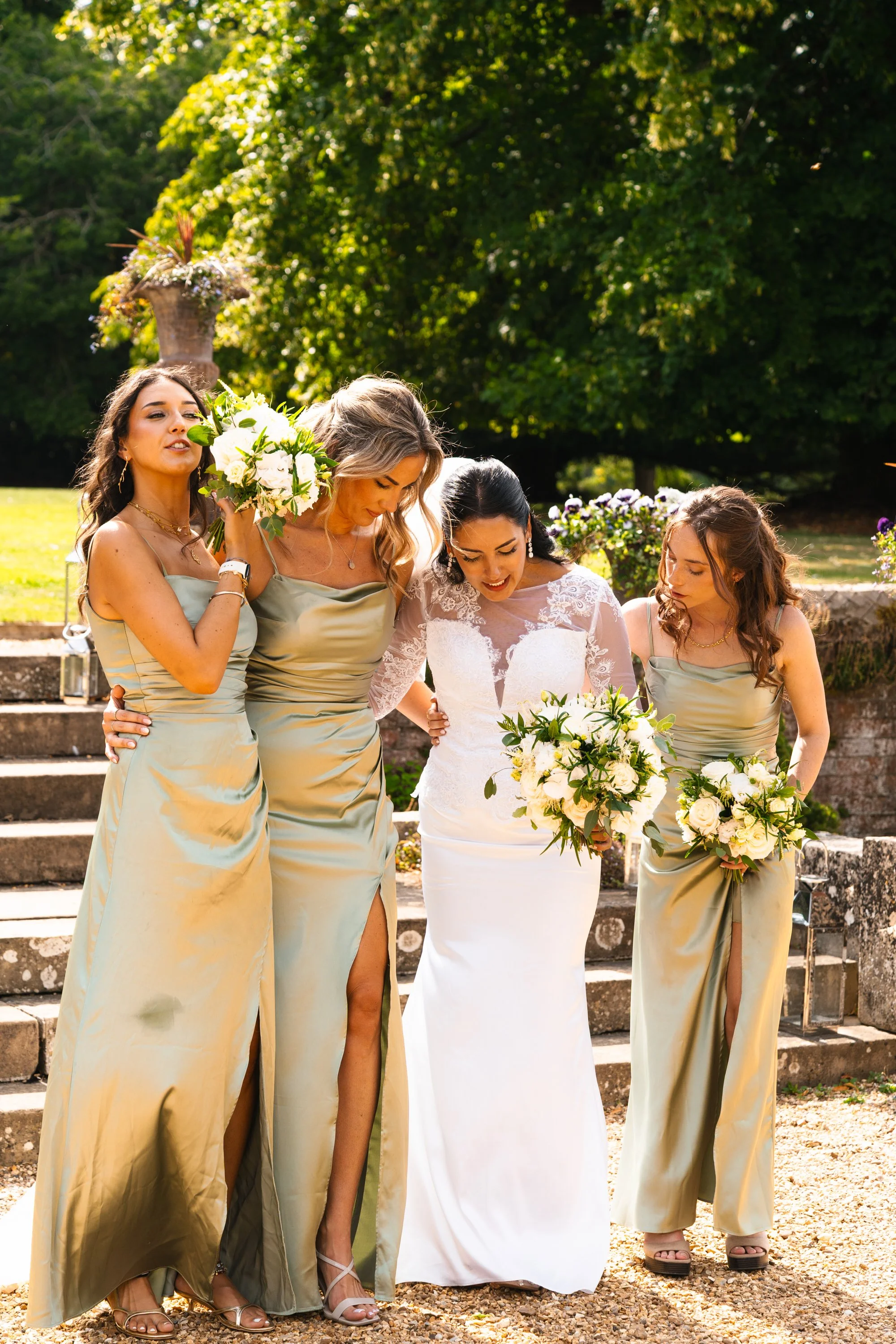 Four women, one dressed in a white wedding gown and three in matching champagne-colored bridesmaid dresses, holding bouquets, walking together outdoors on stone stairs with greenery in the background.