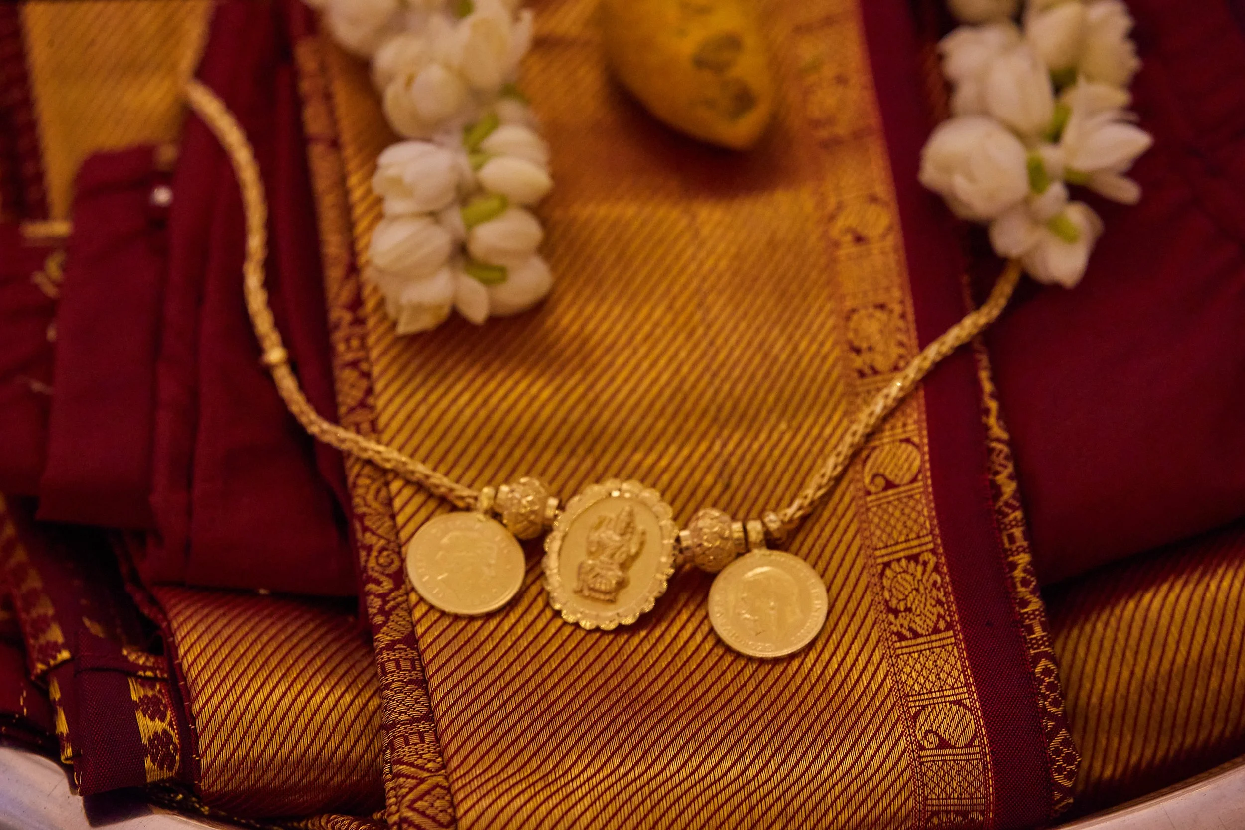 A gold necklace with three medallions rests on a decorative cloth, surrounded by white flowers.