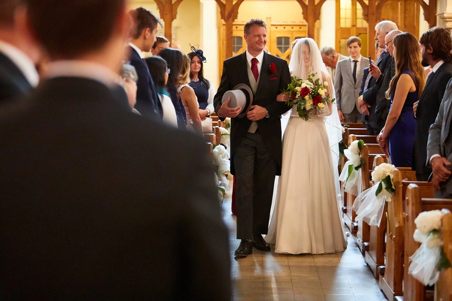 A bride and a man, likely her father, walk down the aisle of a church during a wedding ceremony. The bride is holding a bouquet of flowers and wearing a white wedding dress and veil, while the man is dressed in a suit and holding a gray hat.