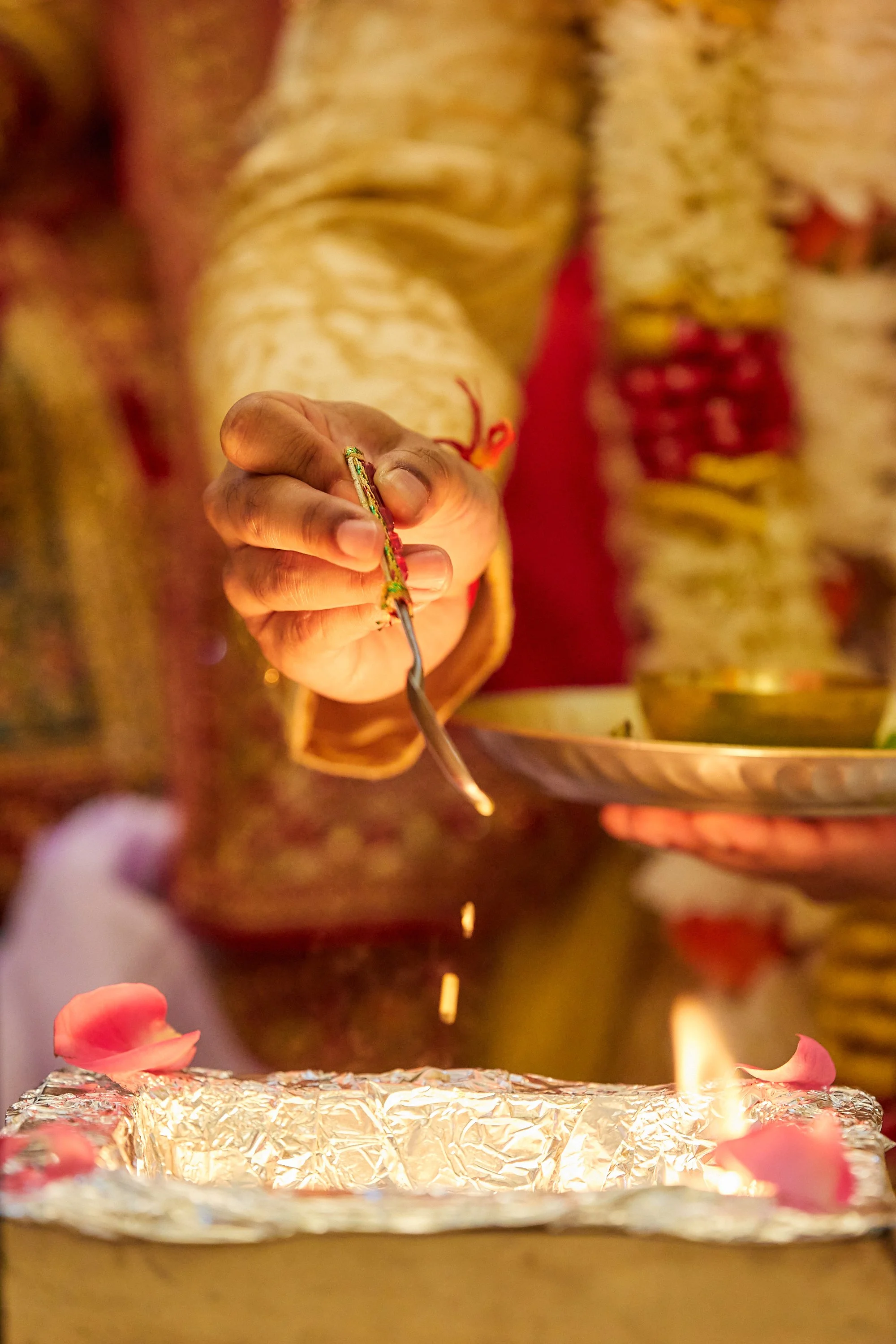A person dressed in a jewelry and traditional attire offering a ritual or prayer, holding a spoon with glowing or burning substance, with flower petals and a lit lamp in the foreground.