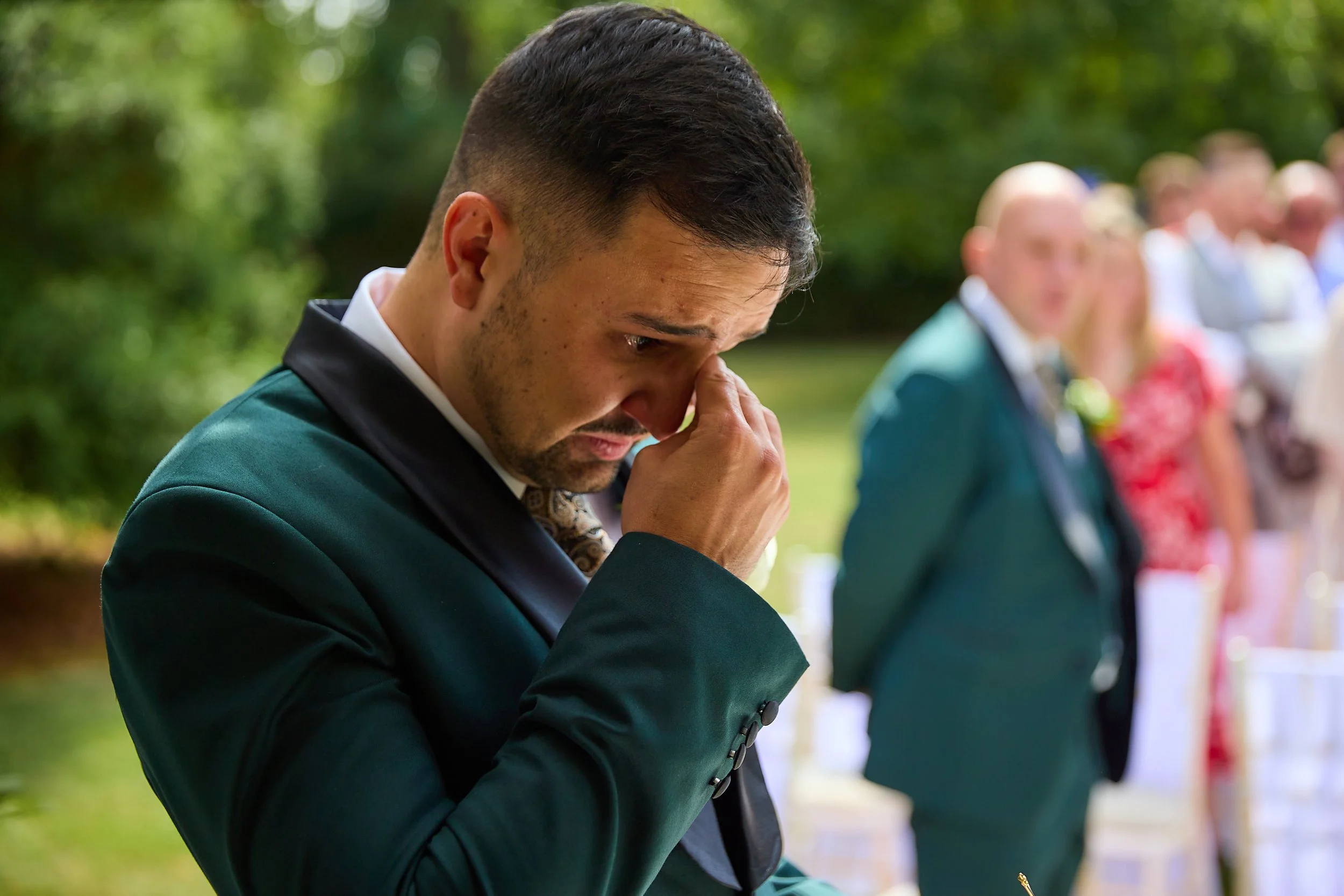 A man dressed in a suit appears to be emotional, gently touching his eye while standing outdoors at a gathering with other people in the background.