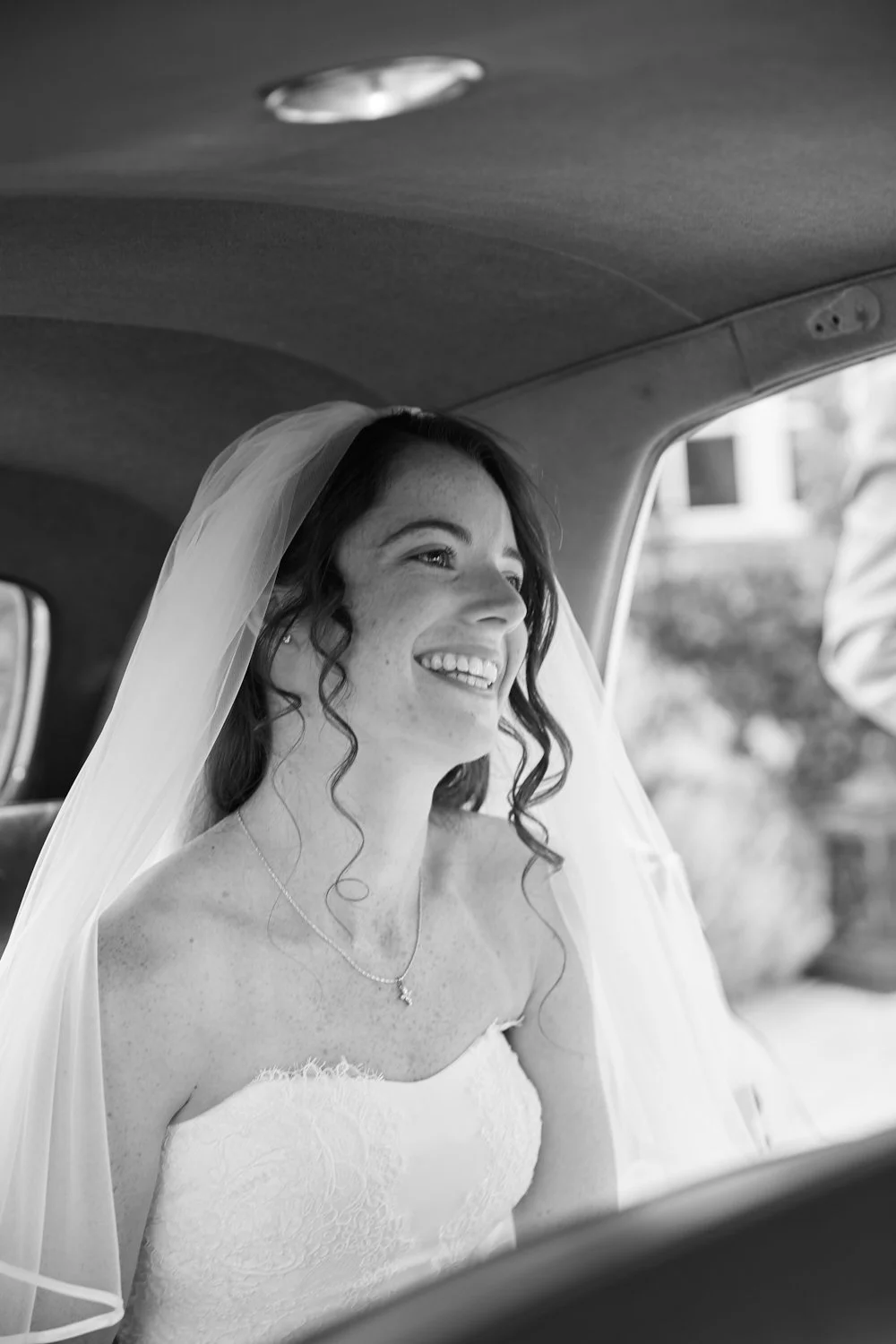 Black and white photo of a smiling bride with a veil sitting inside a vehicle, looking out the window.