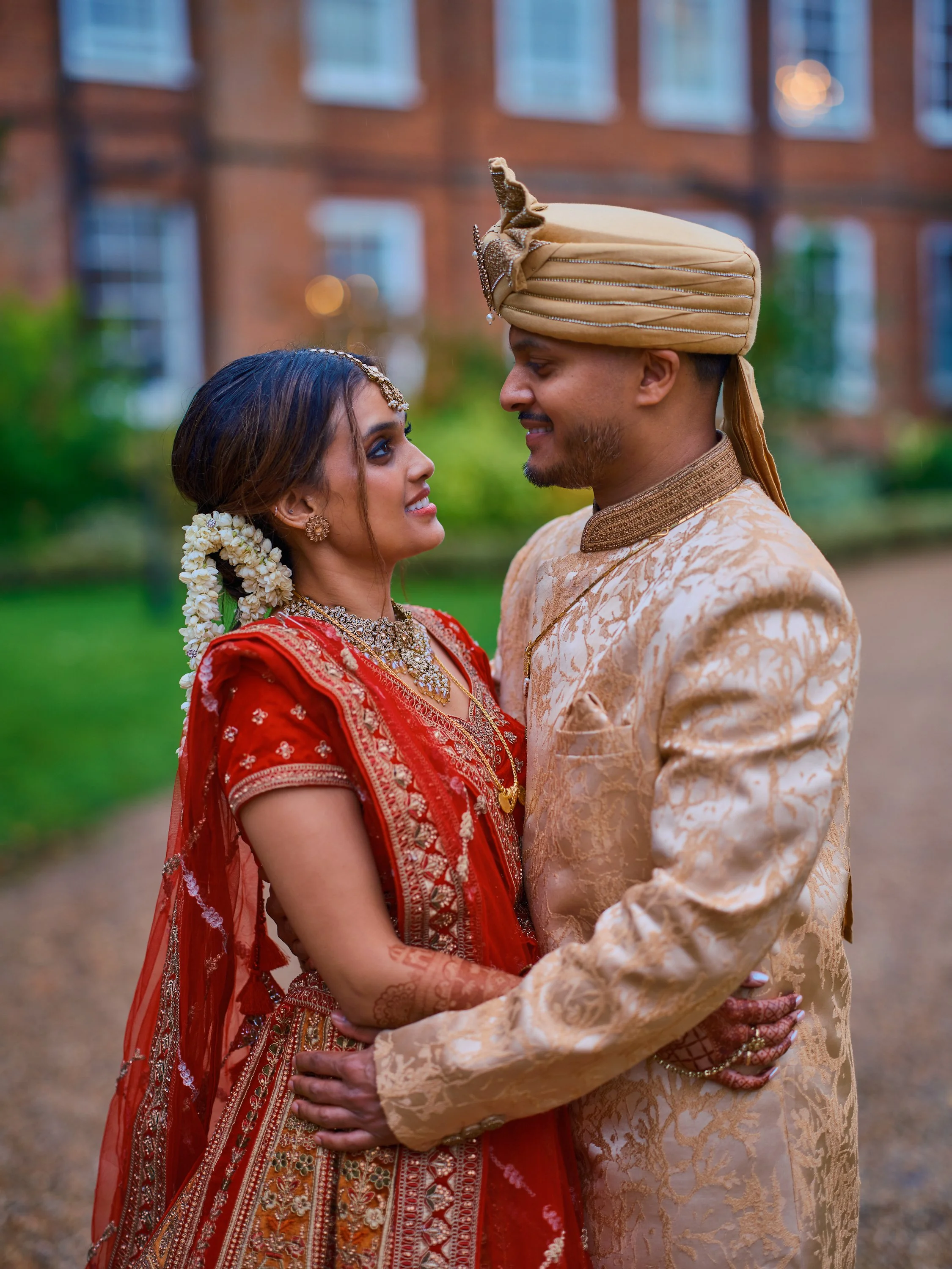 A couple dressed in traditional Indian wedding attire, standing outdoors and gazing into each other's eyes.