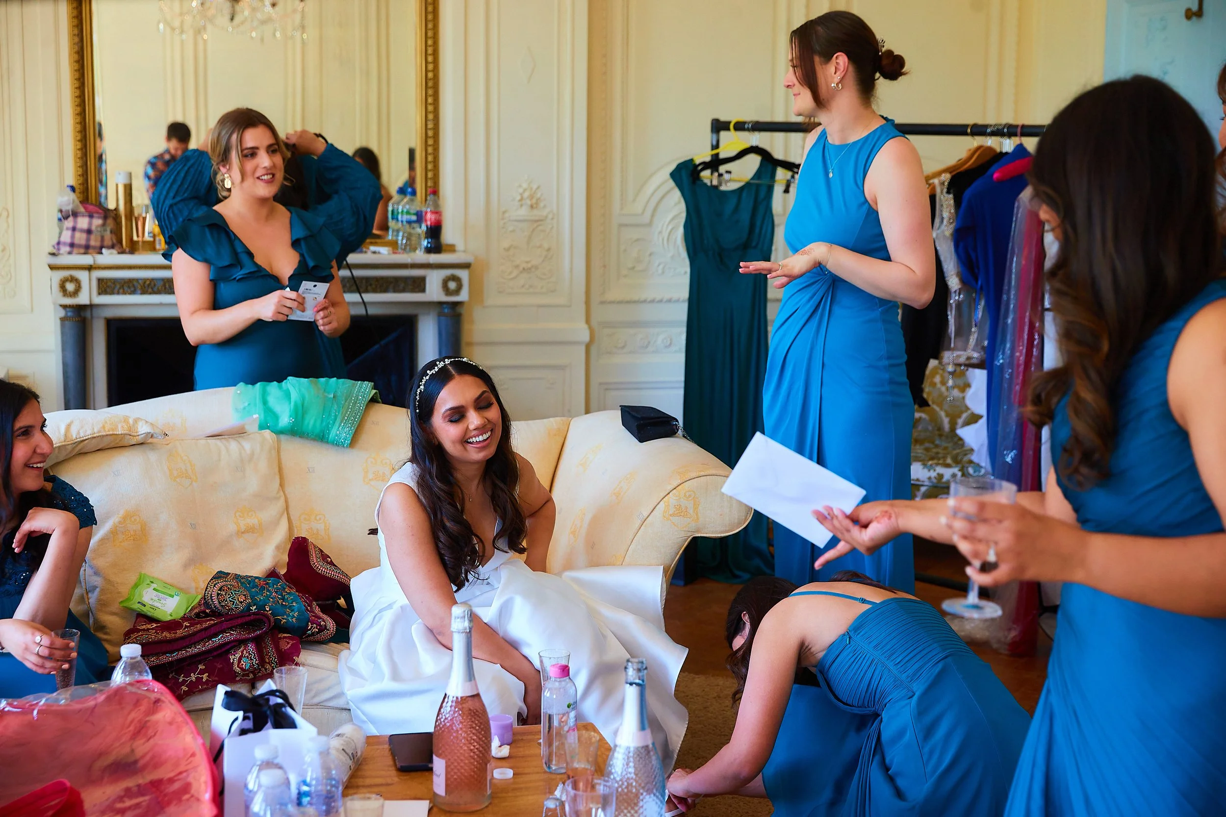 A bride sitting on a sofa surrounded by her bridesmaids preparing for her wedding, with some sitting, some standing, and one leaning forward on the floor, in an elegant room with a large mirror and decorated walls.