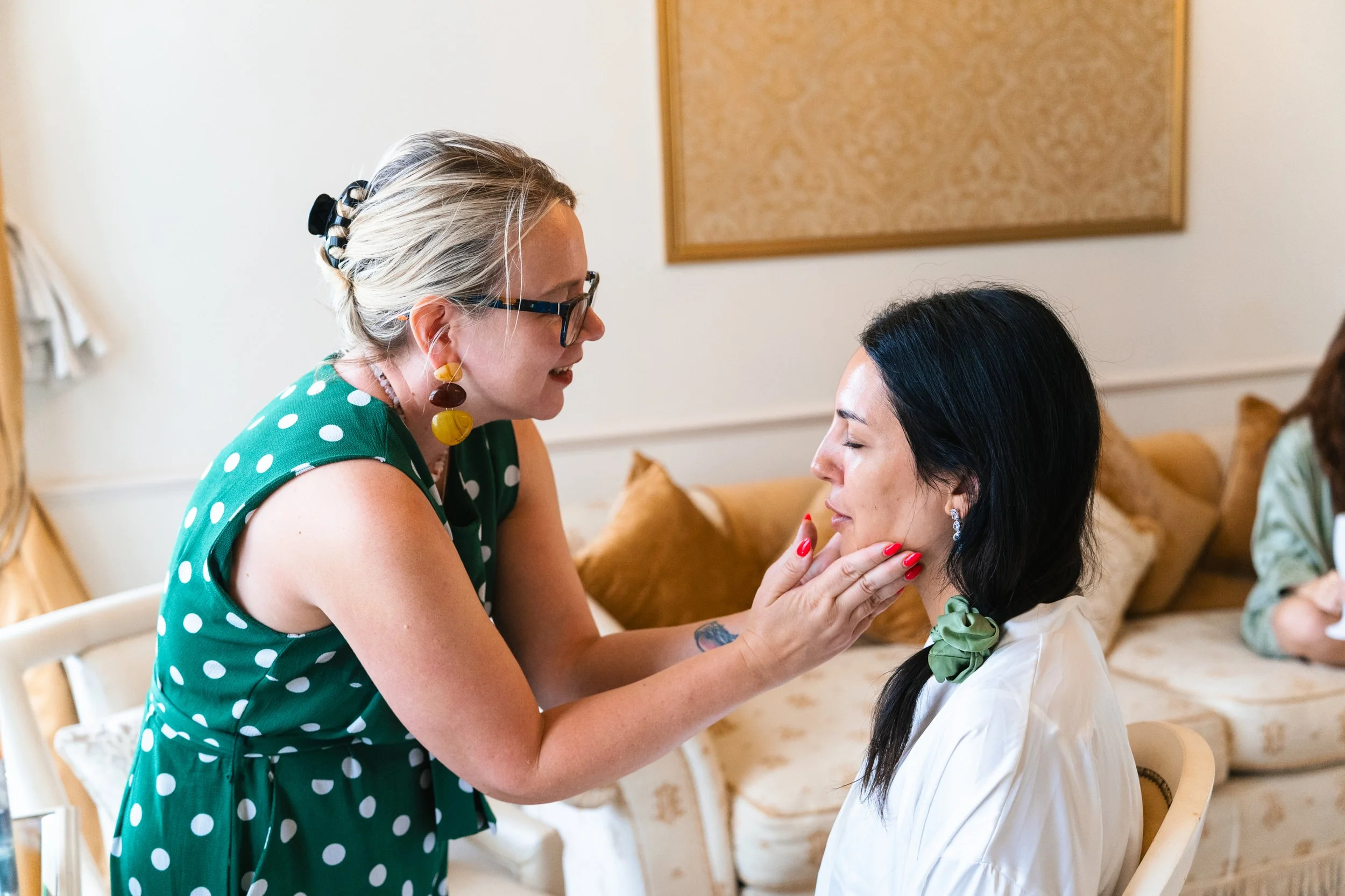 Woman with glasses and earrings gently touches another woman's chin with her hand in a cozy, well-lit room.